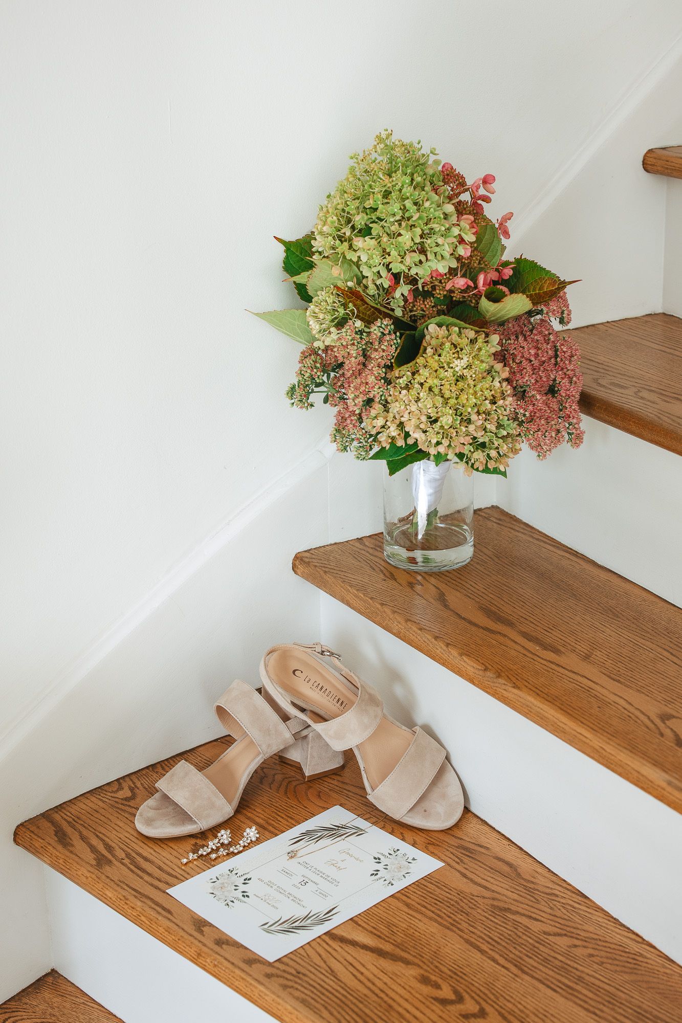 Sandales beige et carte d'invitation pour un mariage sur des marches en bois, à côté d’un vase de fleurs avec hortensias et fleurs roses