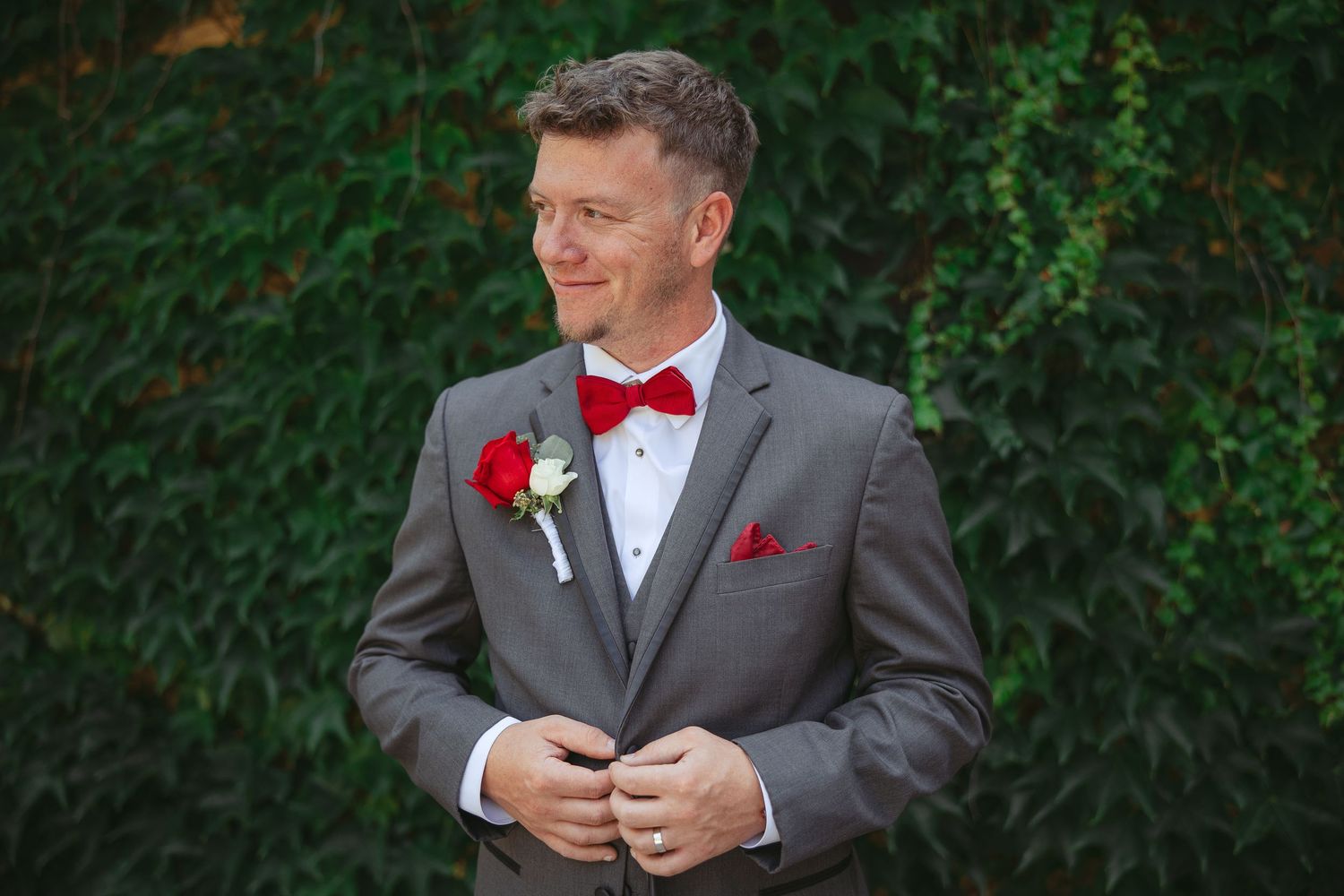 A groom in a grey suit with red bow tie and boutonniere smiles while adjusting his jacket.