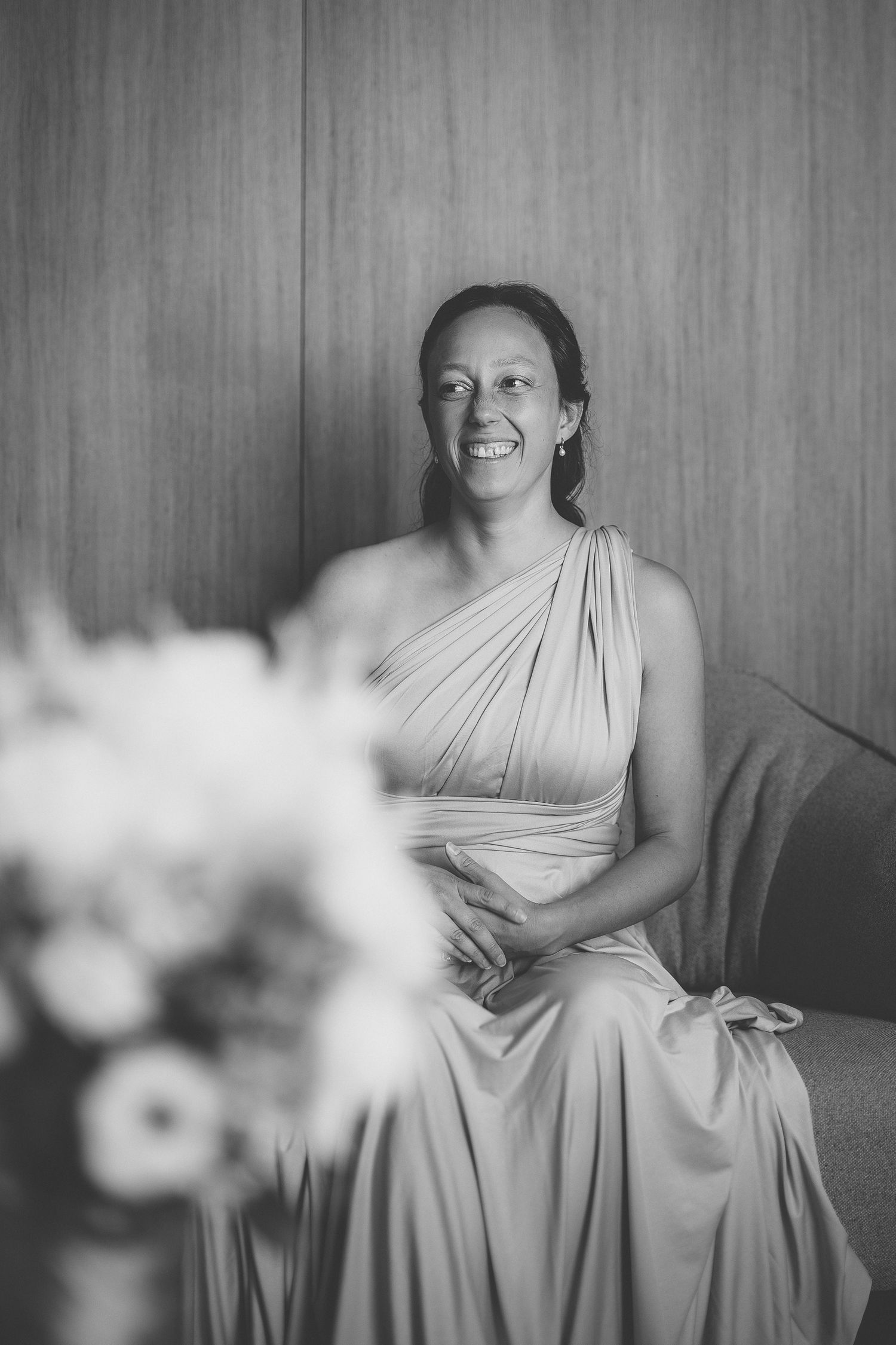 Portraits de mariage en noir et blanc d’une demoiselle d’honneur en robe claire, assise contre un mur de bois lors des préparatifs.