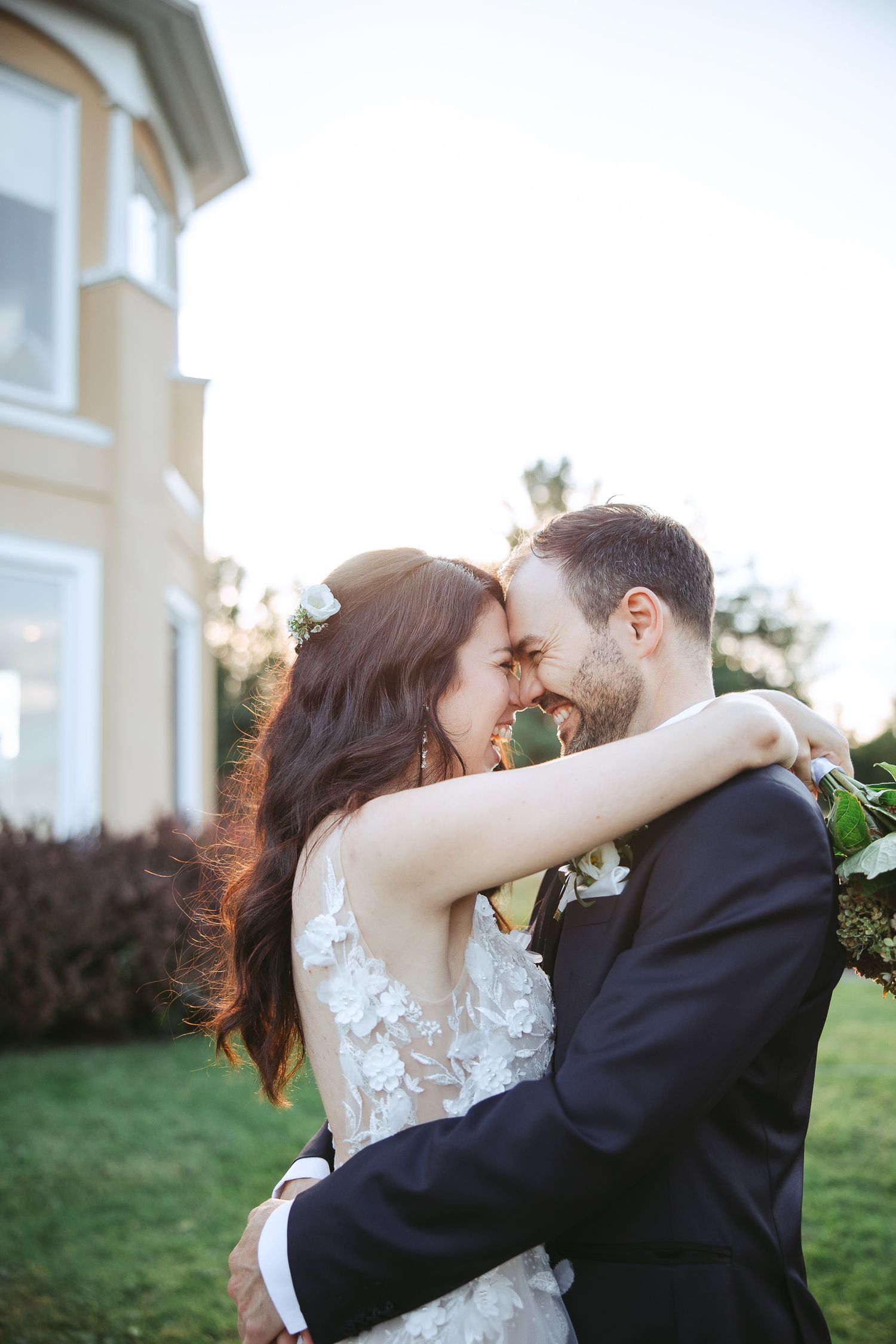 A romantic moment between newlyweds embracing outdoors at sunset near an elegant house with a manicured lawn.