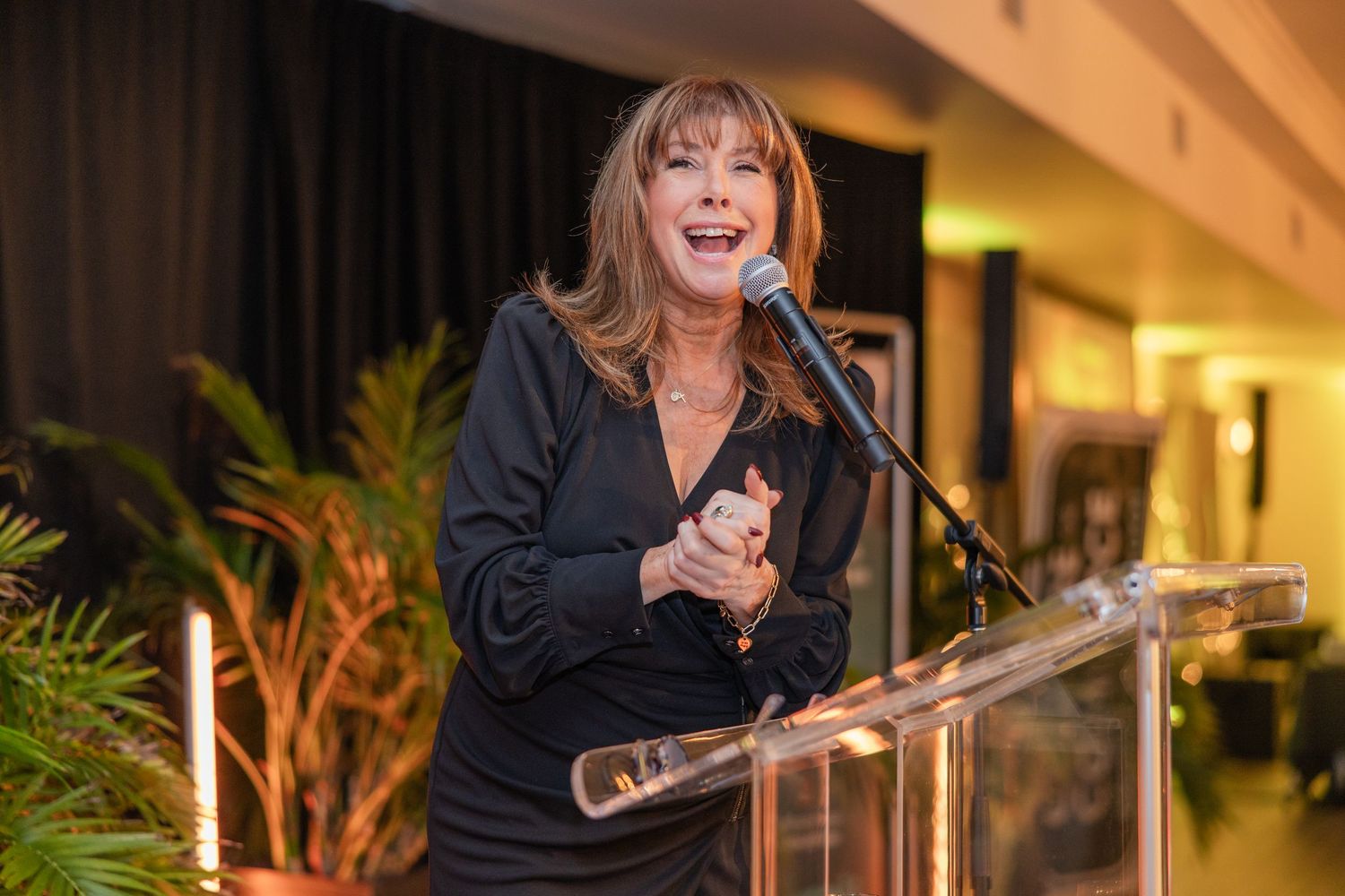 A speaker presents at a podium during an evening event in a warmly lit venue with indoor plants visible.