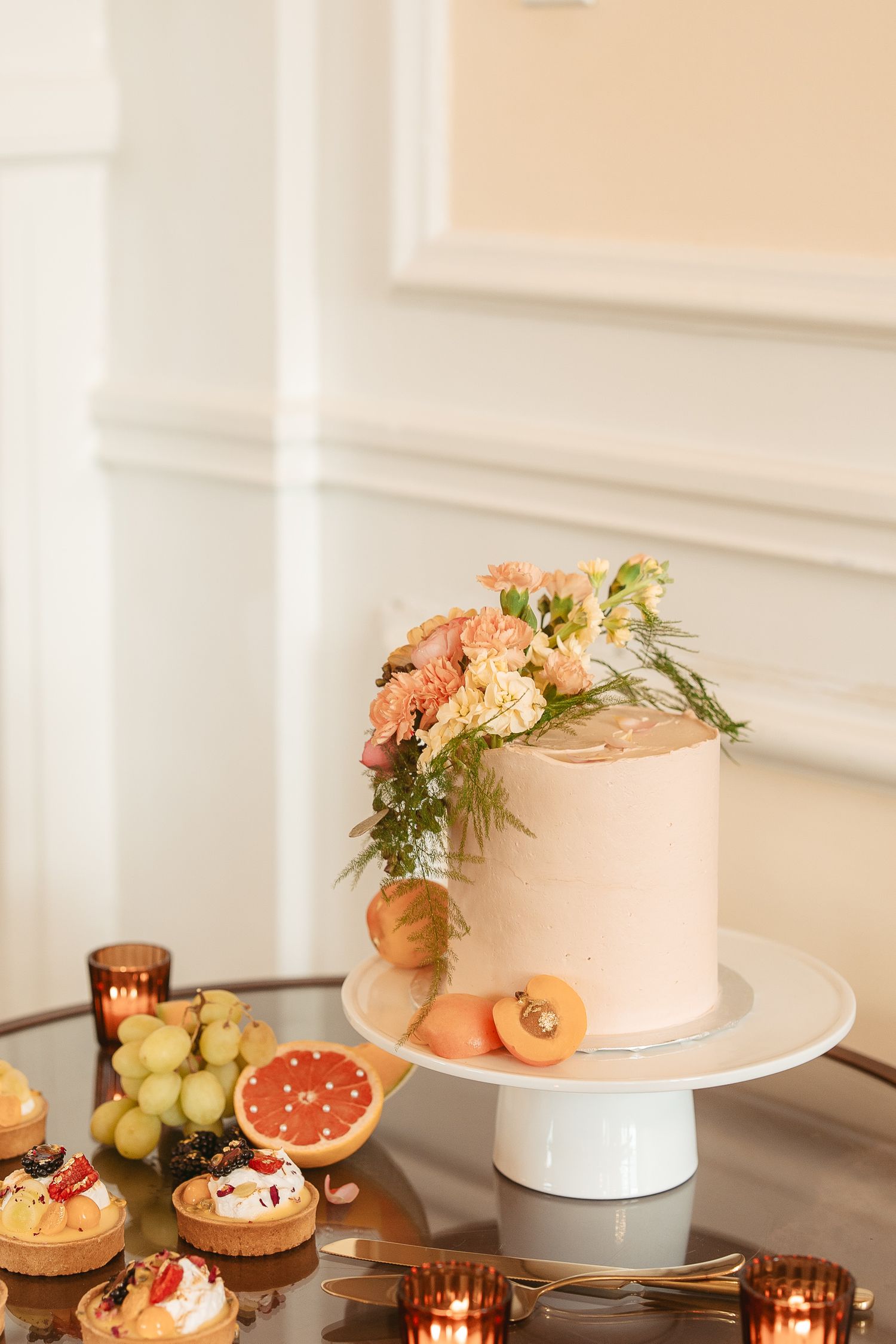 Pink cake with floral decorations surrounded by copper candle holders and fresh fruit on an elegant white table setting.