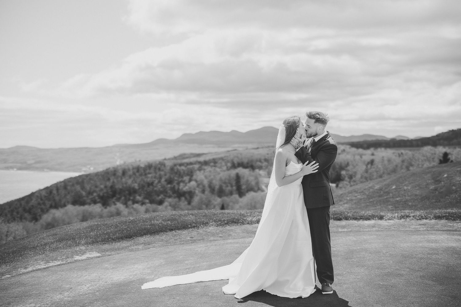Un couple en tenue de mariage s’enlace en se tenant dans un paysage montagneux au coucher du soleil.