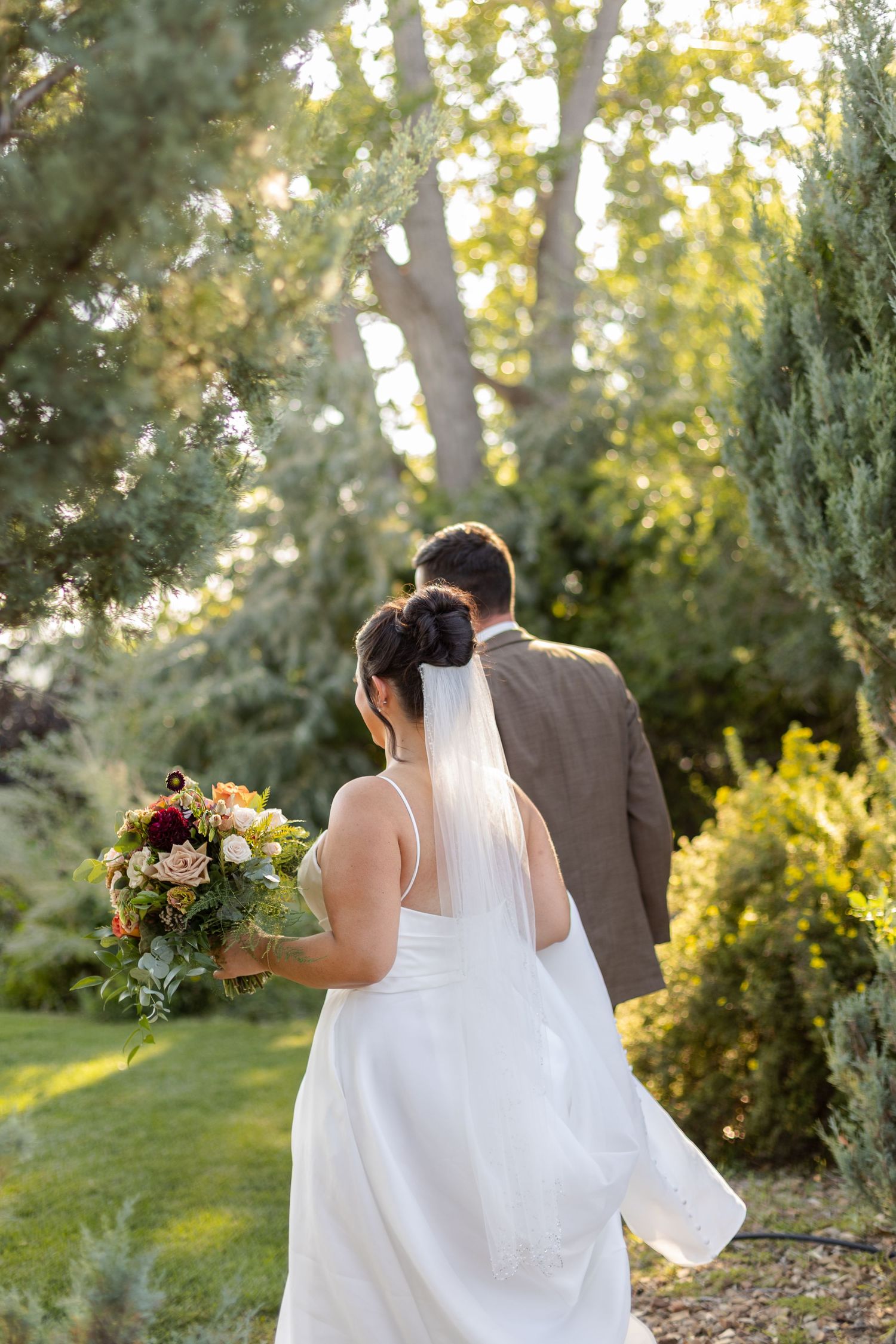 A couple walks together in a garden setting during golden hour sunlight, with the bride holding a floral bouquet.