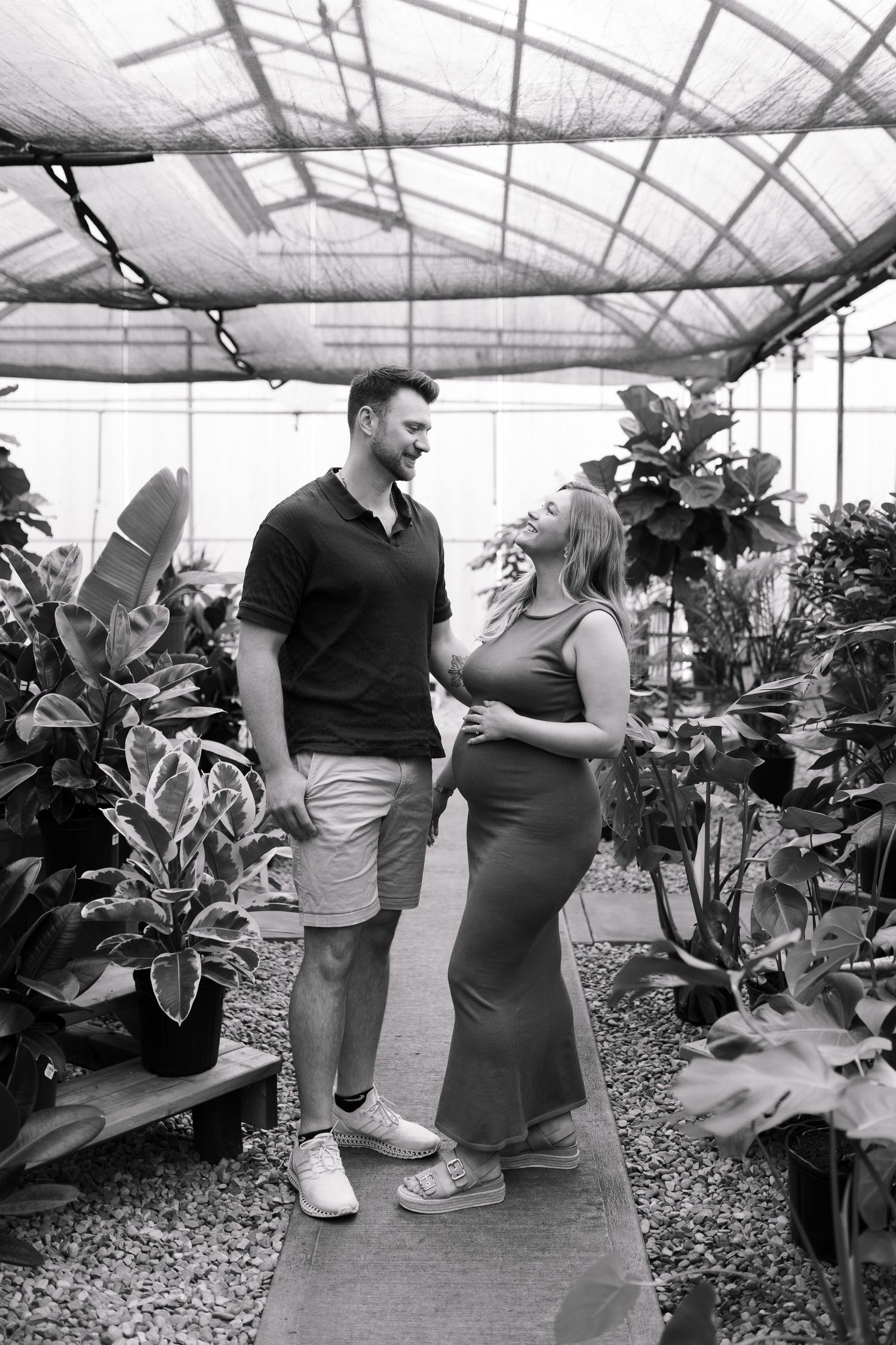 Black and white maternity photo of couple in greenhouse surrounded by tropical plants.