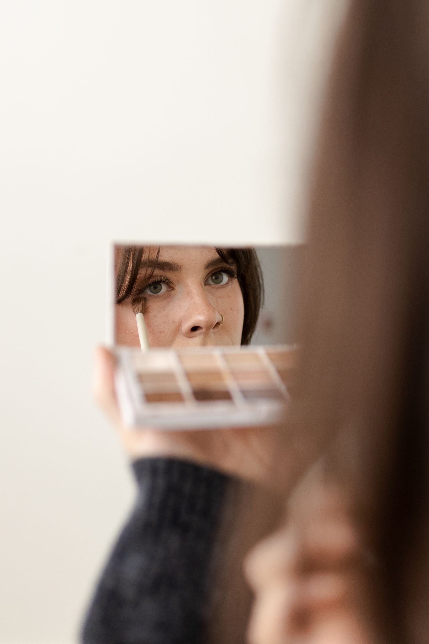 A reflection in a mirror shows someone looking at a makeup palette in a soft, artistic composition.
