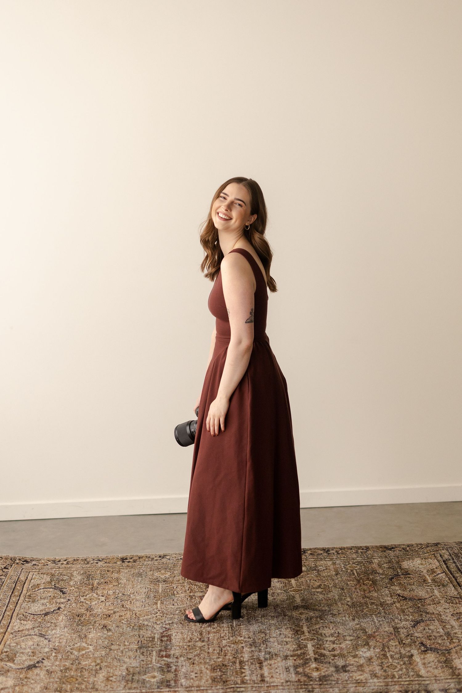 A person in a burgundy sleeveless dress poses against a white wall on a textured vintage rug.