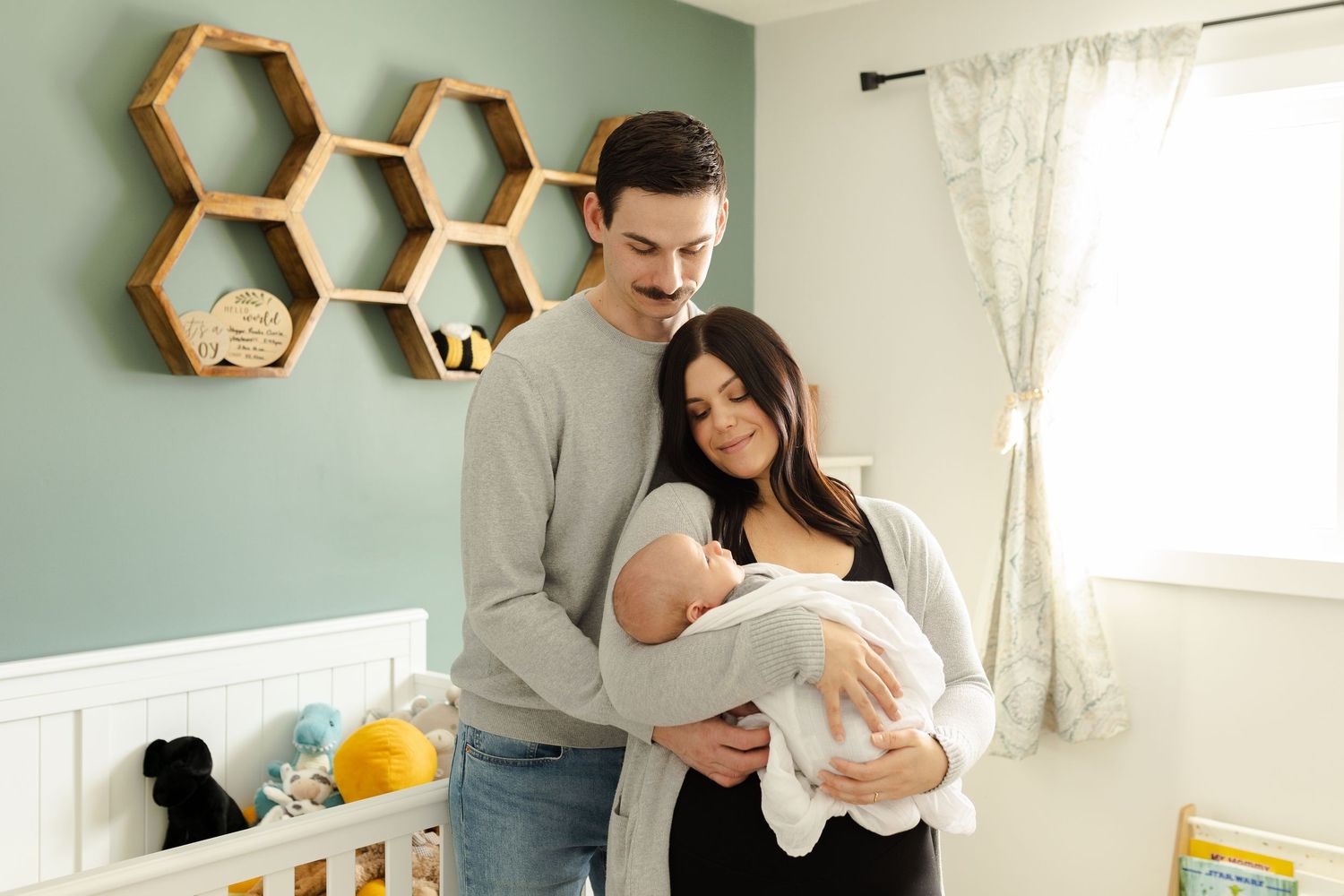 A couple stands together in a nursery room with honeycomb-shaped shelves and mint green walls.