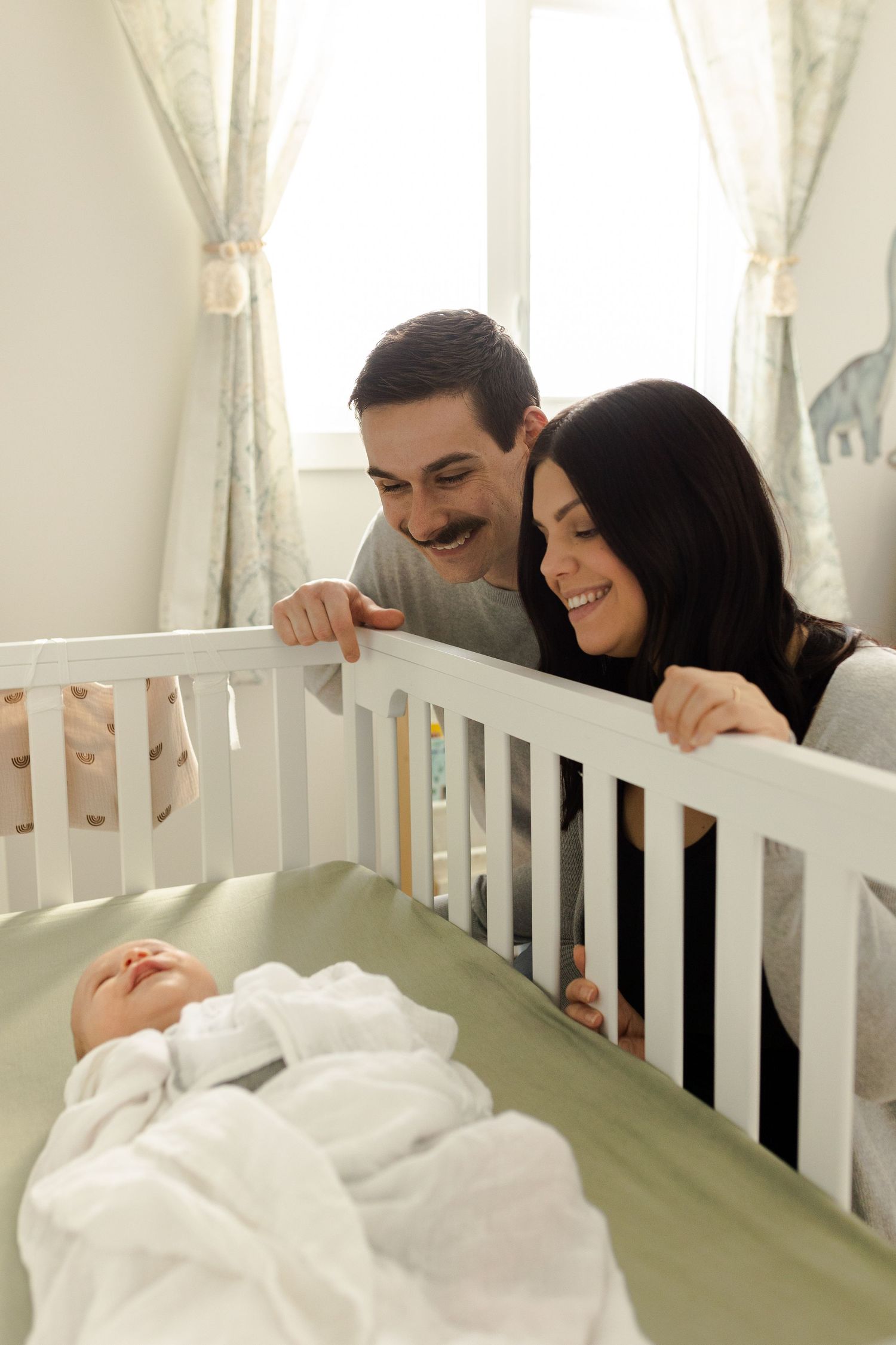A couple peeks over the edge of a white crib together in a nursery setting.