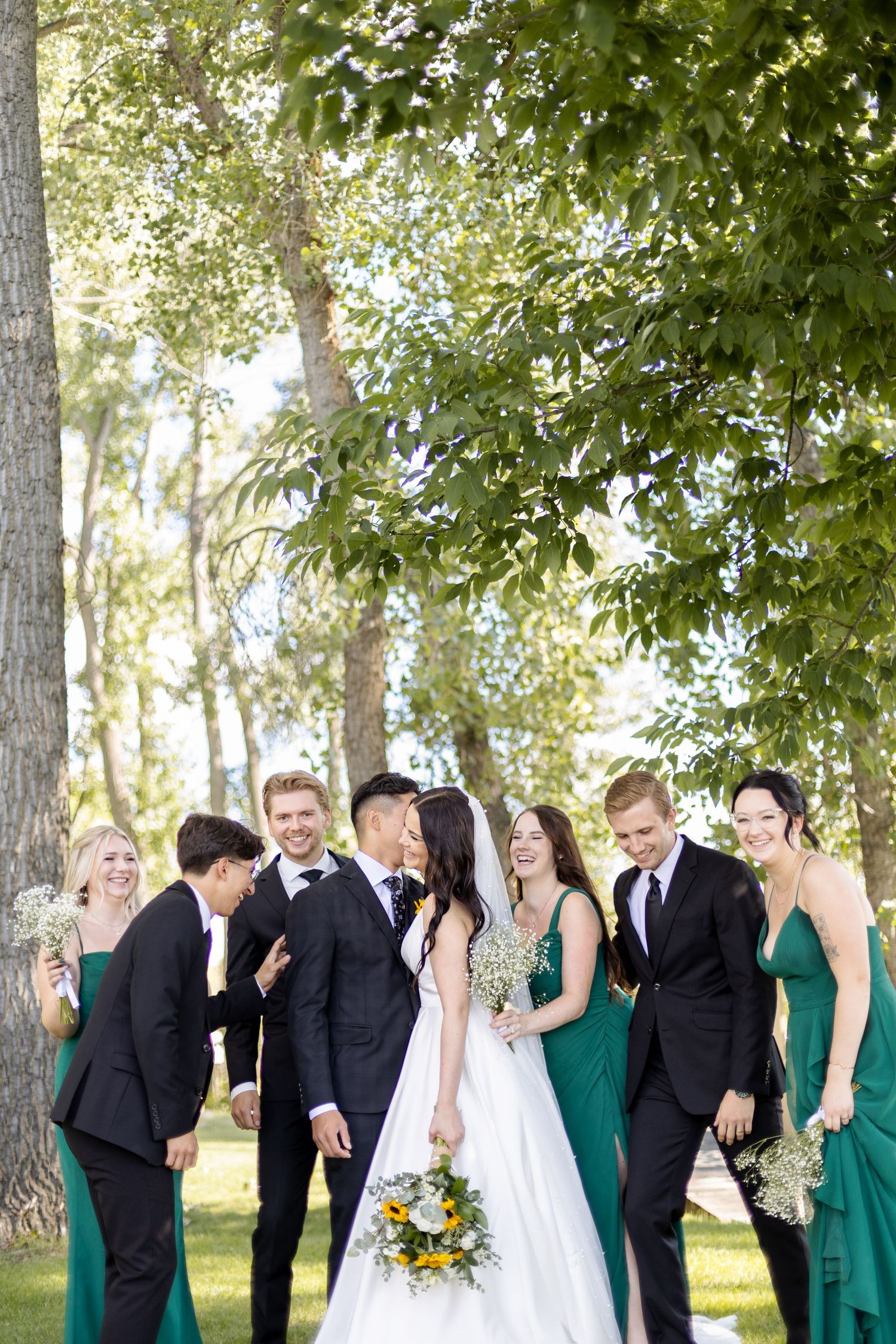 Wedding party in black suits and emerald green dresses gathered under trees for a candid group photo.