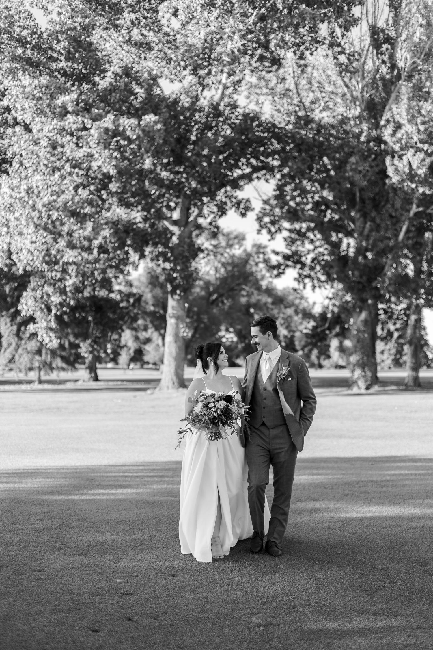 Black and white photo of couple walking under large trees in park setting.