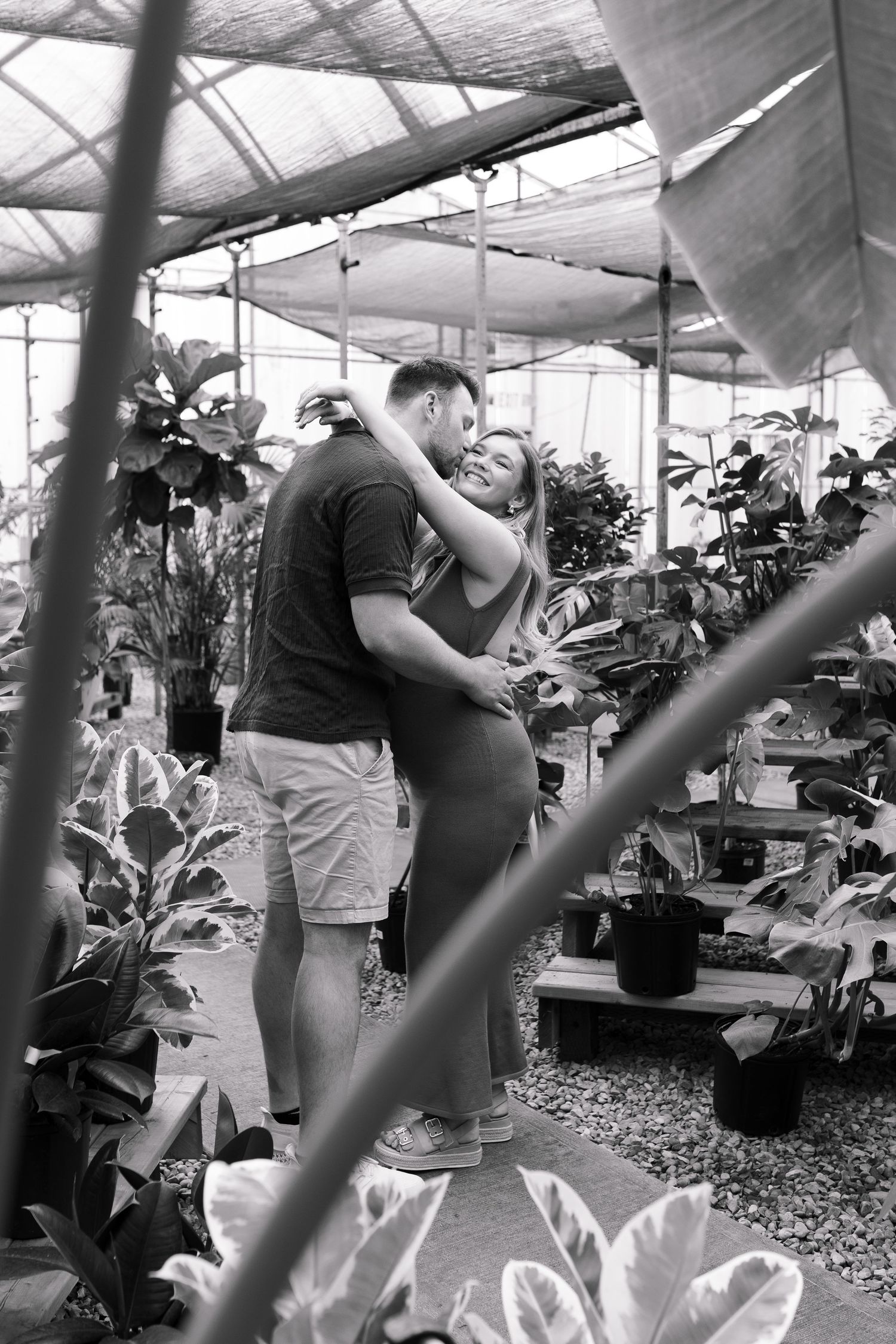 Black and white romantic greenhouse photo with couple embracing among potted plants.