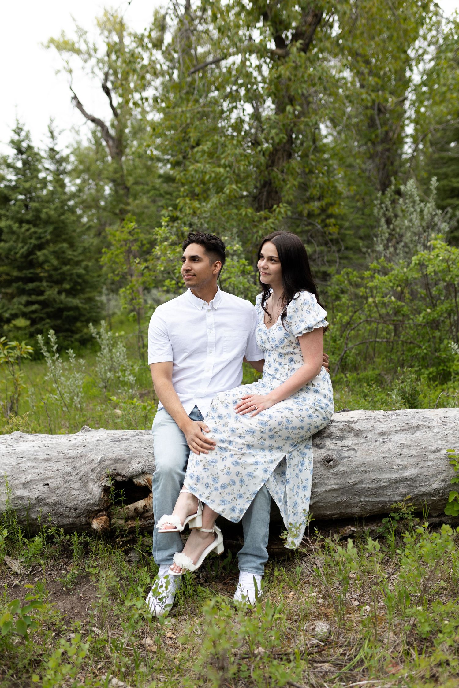 A couple sits together on a fallen tree trunk in a forest setting wearing coordinated outfits.