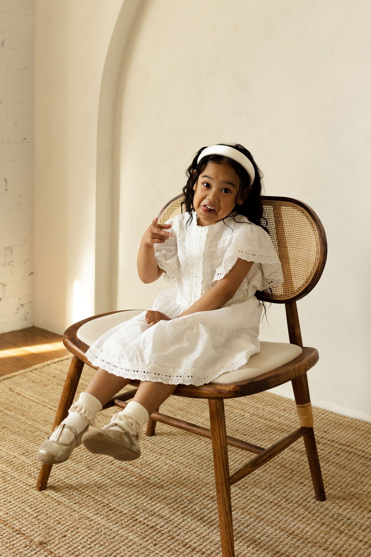 Child in white dress playing on vintage wooden chair against neutral background.
