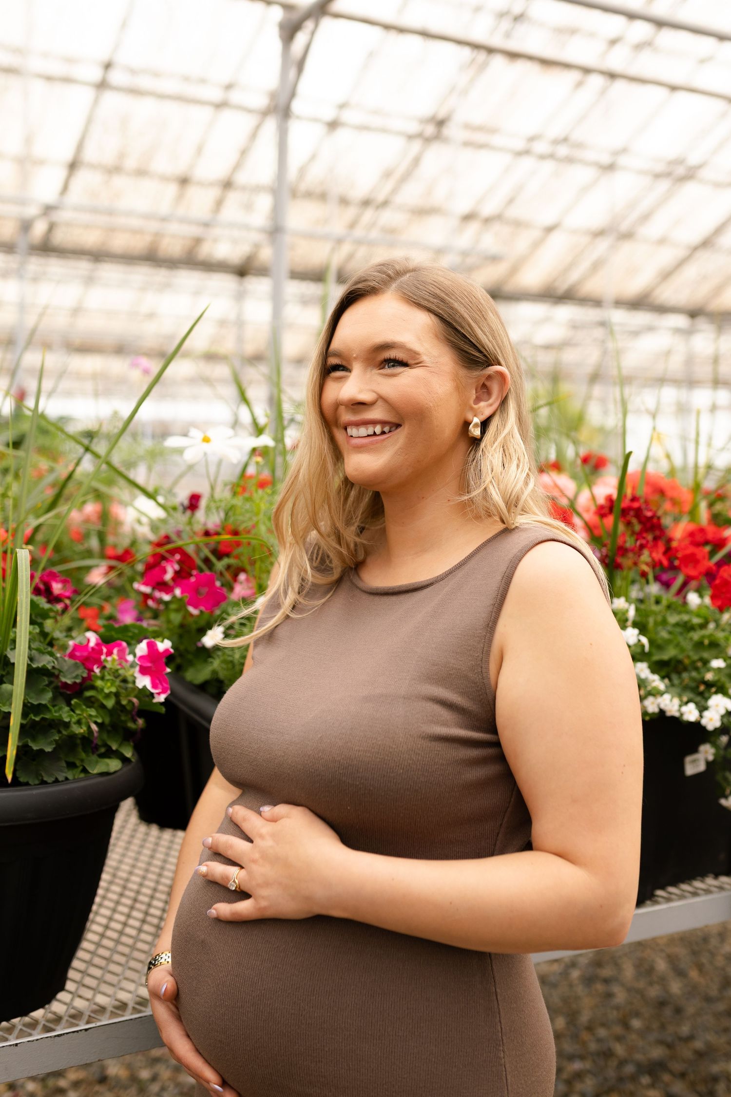 Expecting mother in brown dress smiles while standing in a greenhouse with colorful flowers.