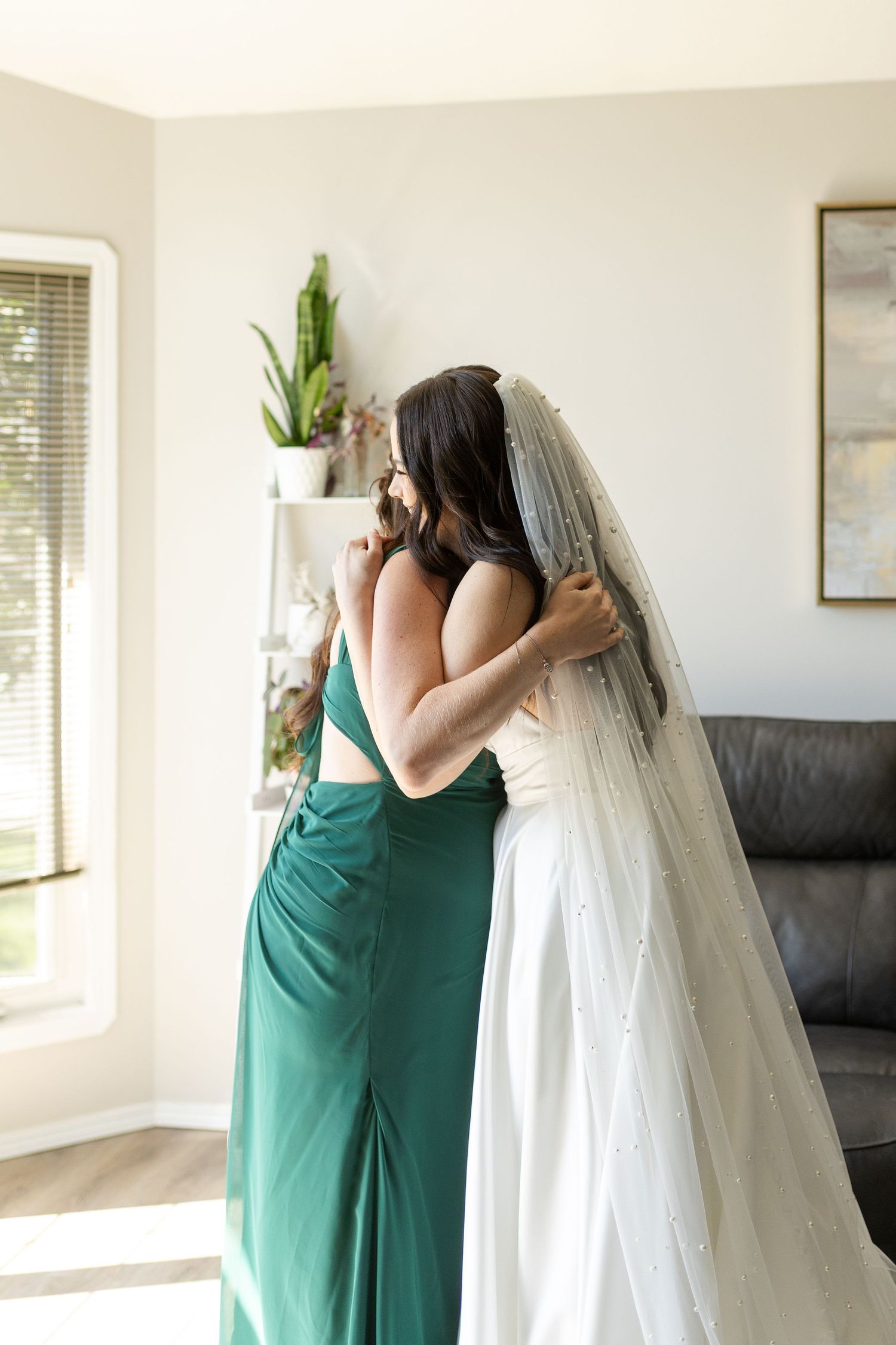Emotional first look moment between bride in white gown and bridesmaid in emerald green dress indoors.