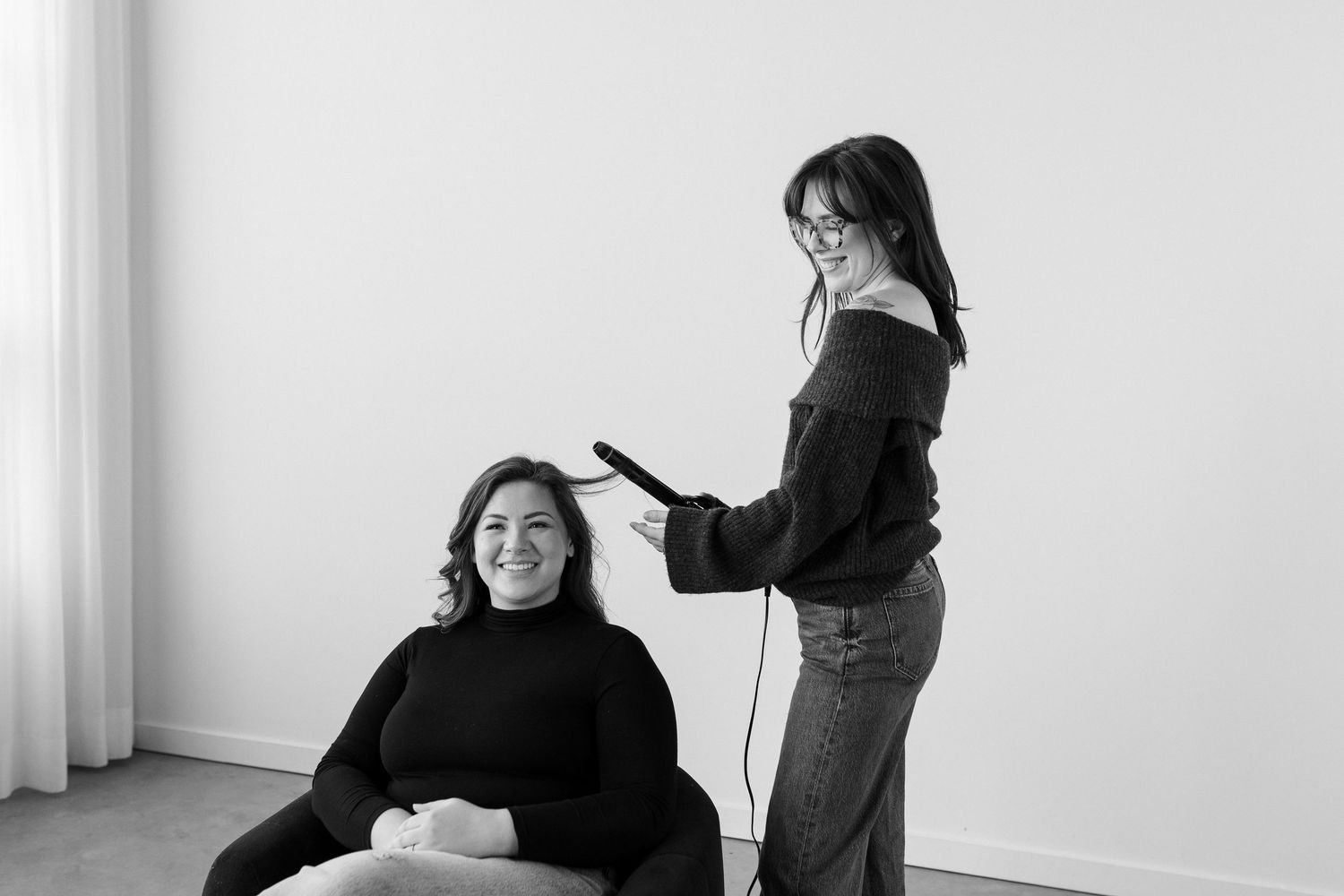 A stylist uses a flat iron while working on a client's hair in a minimalist white studio setting.