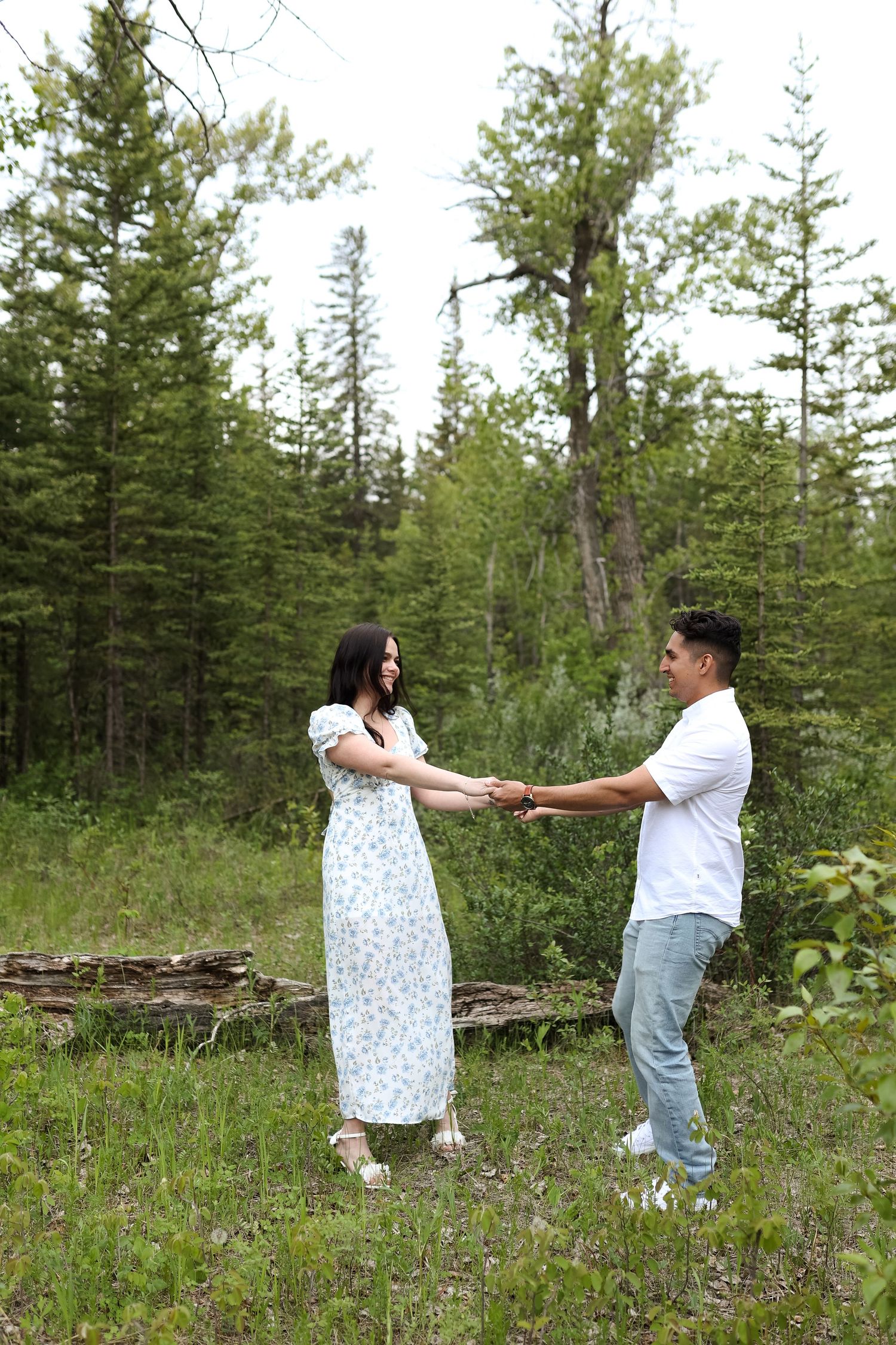 A couple holds hands while dancing in a forest clearing wearing coordinated white outfits.