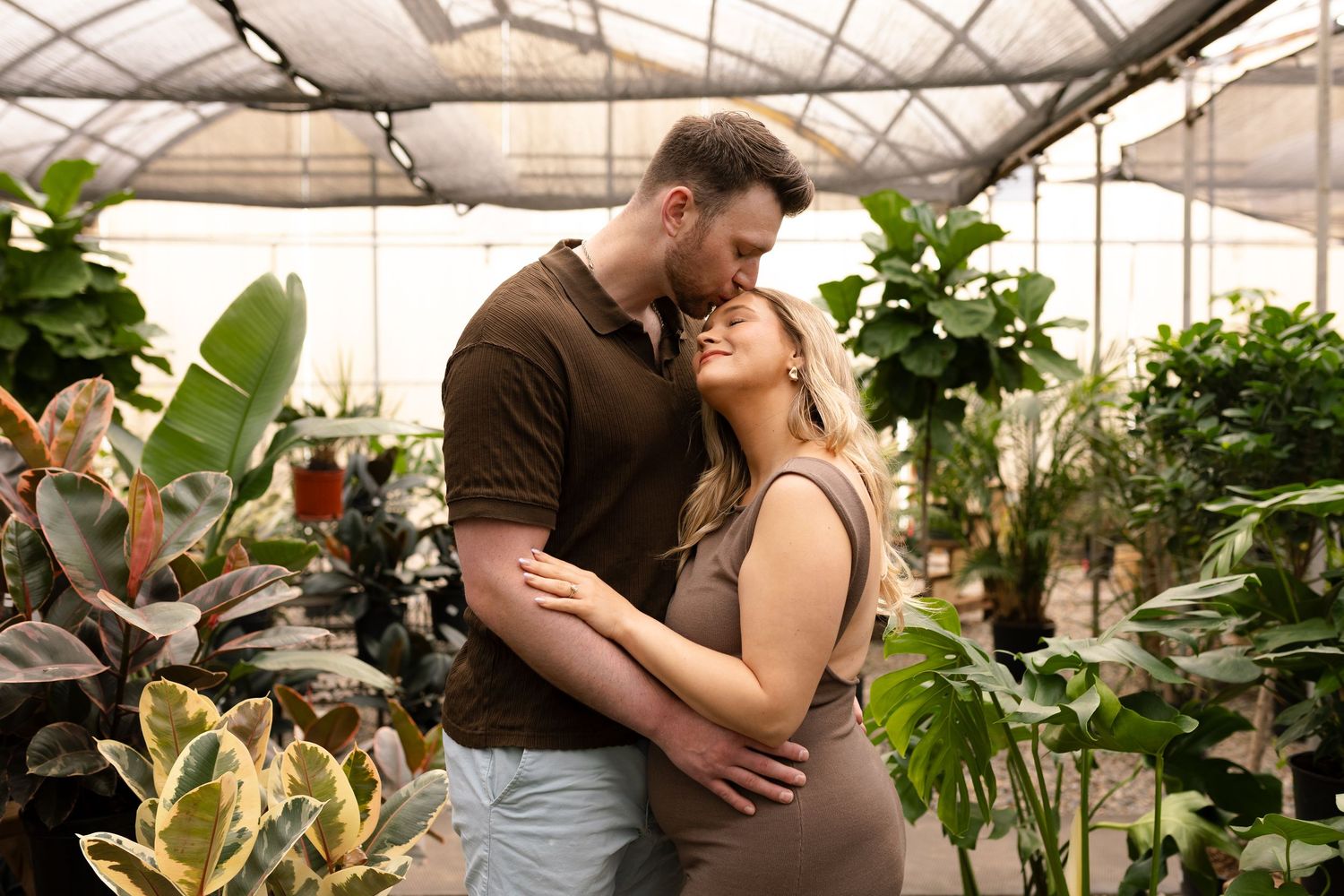 Intimate maternity photo of couple embracing in greenhouse surrounded by tropical foliage.