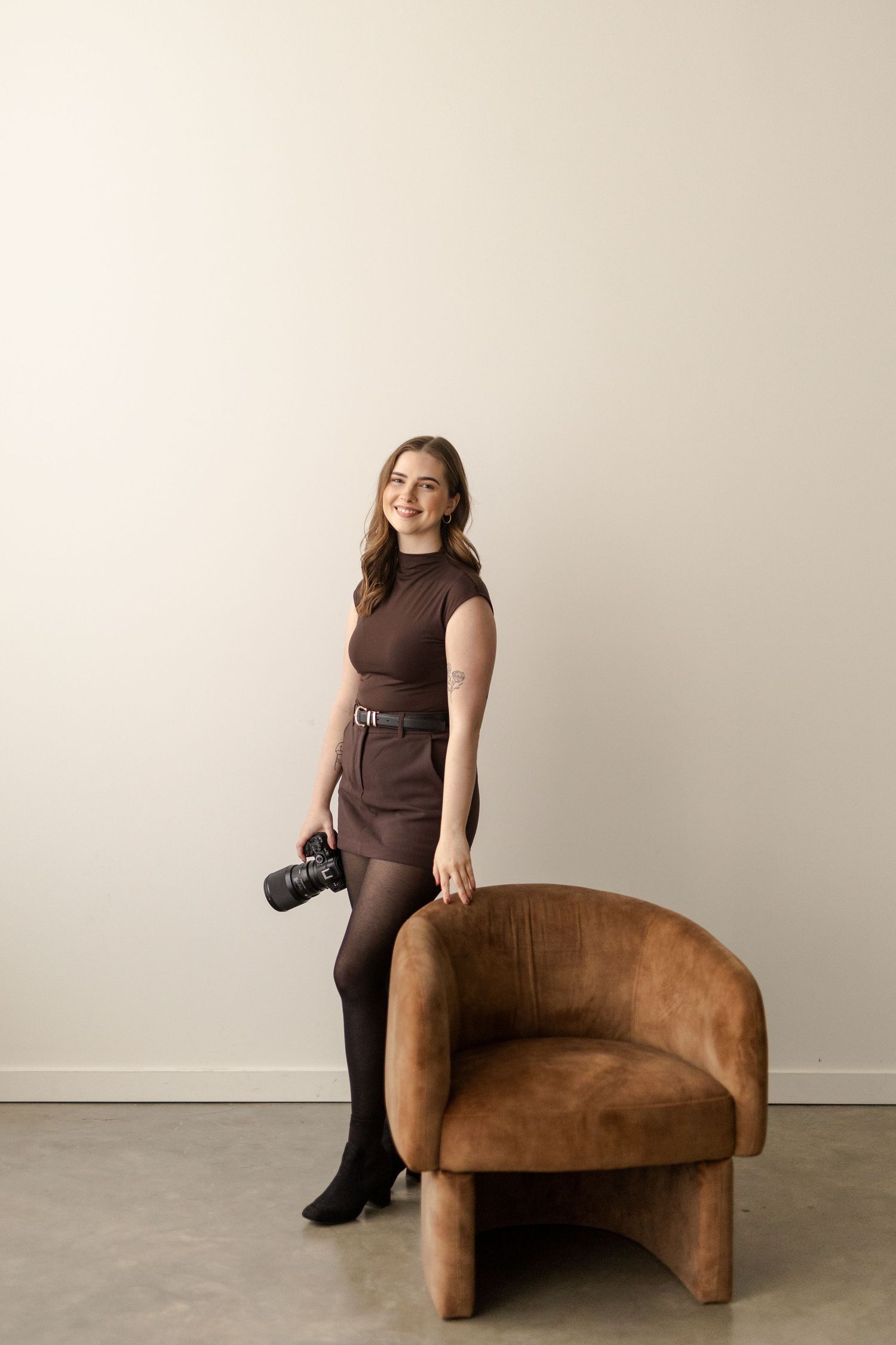 A photographer poses with a brown leather chaise lounge in a minimalist studio setting against a neutral wall.