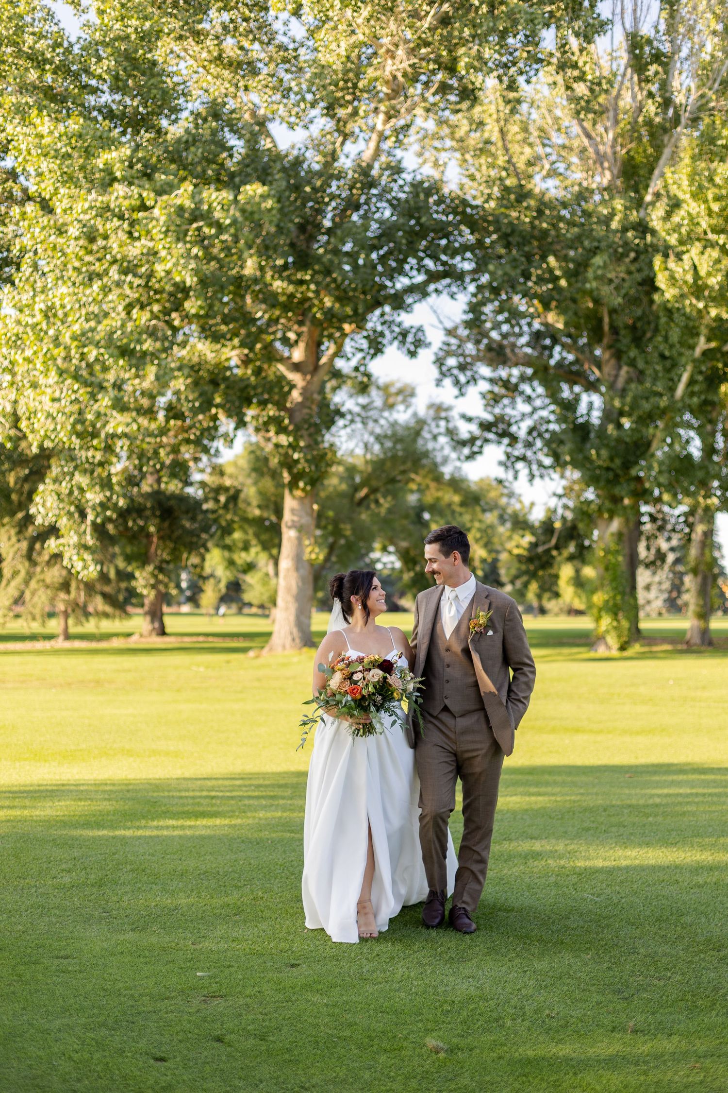 A wedding couple walks hand in hand through a scenic park with tall trees in golden afternoon light.