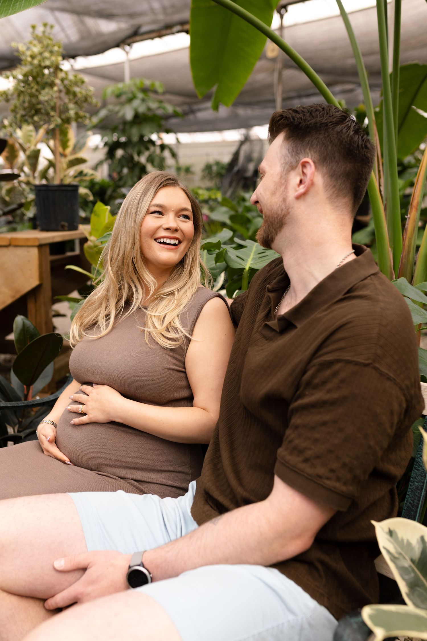 Joyful greenhouse maternity photo with couple laughing together among tropical plants.