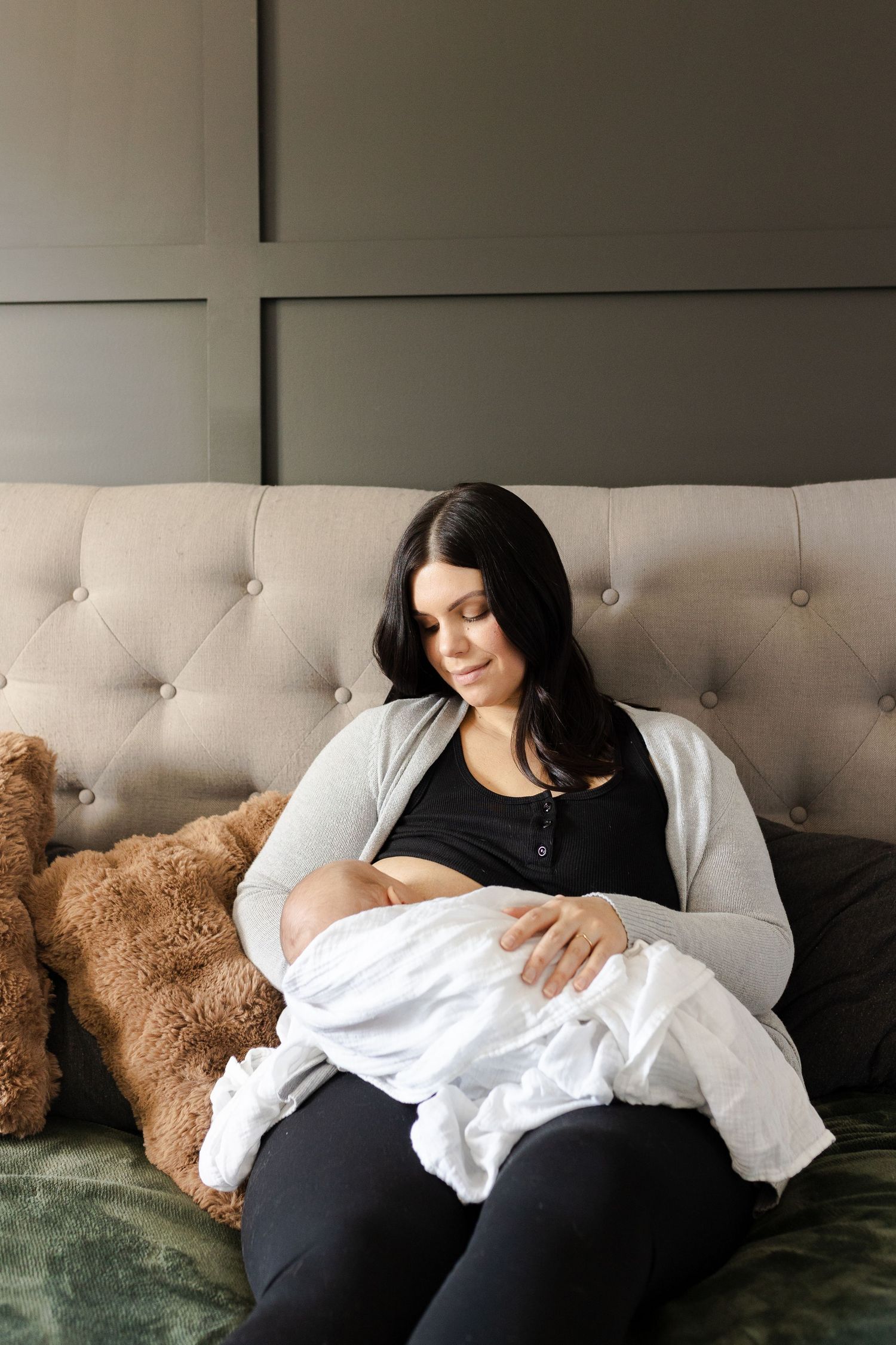 Mother in gray cardigan nursing baby while resting on tufted couch with teddy bear.