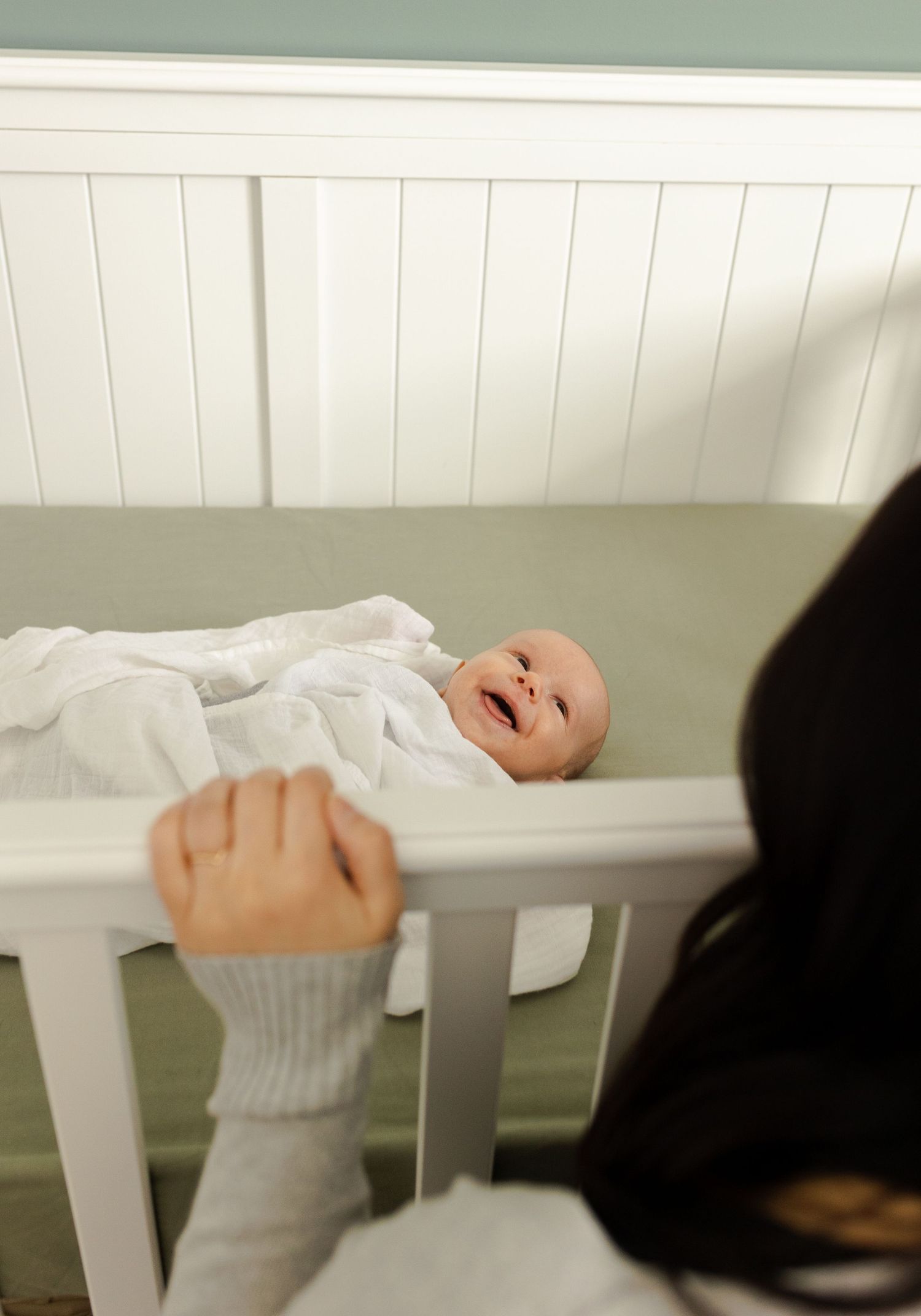 A smiling baby lies in a white crib while being watched from above.