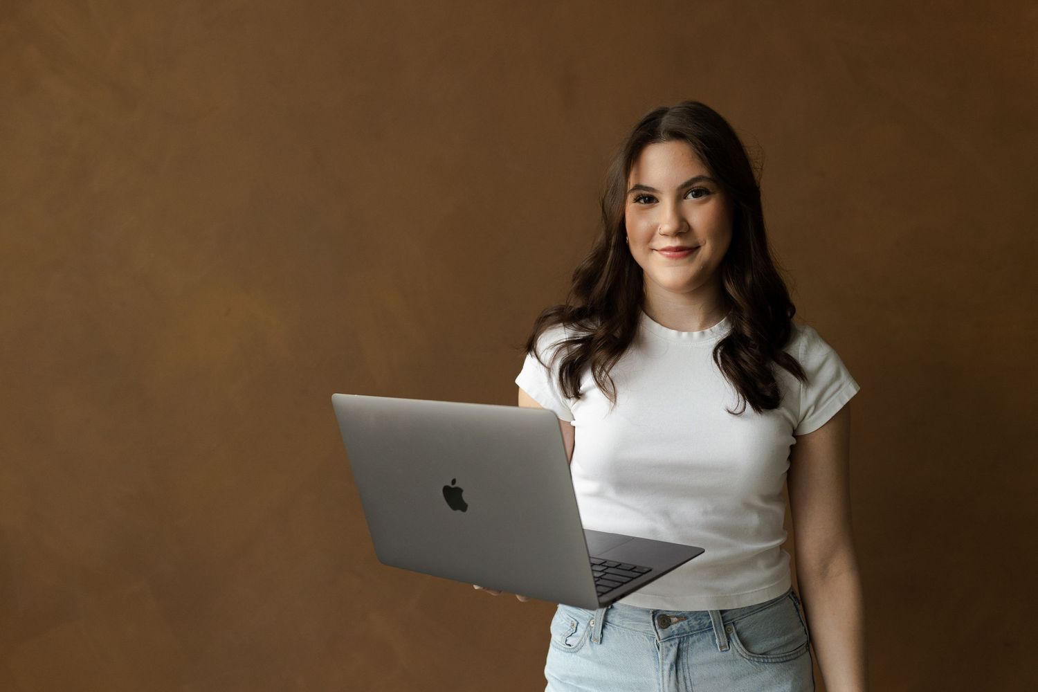 Person in white shirt working on MacBook laptop against brown wall background.