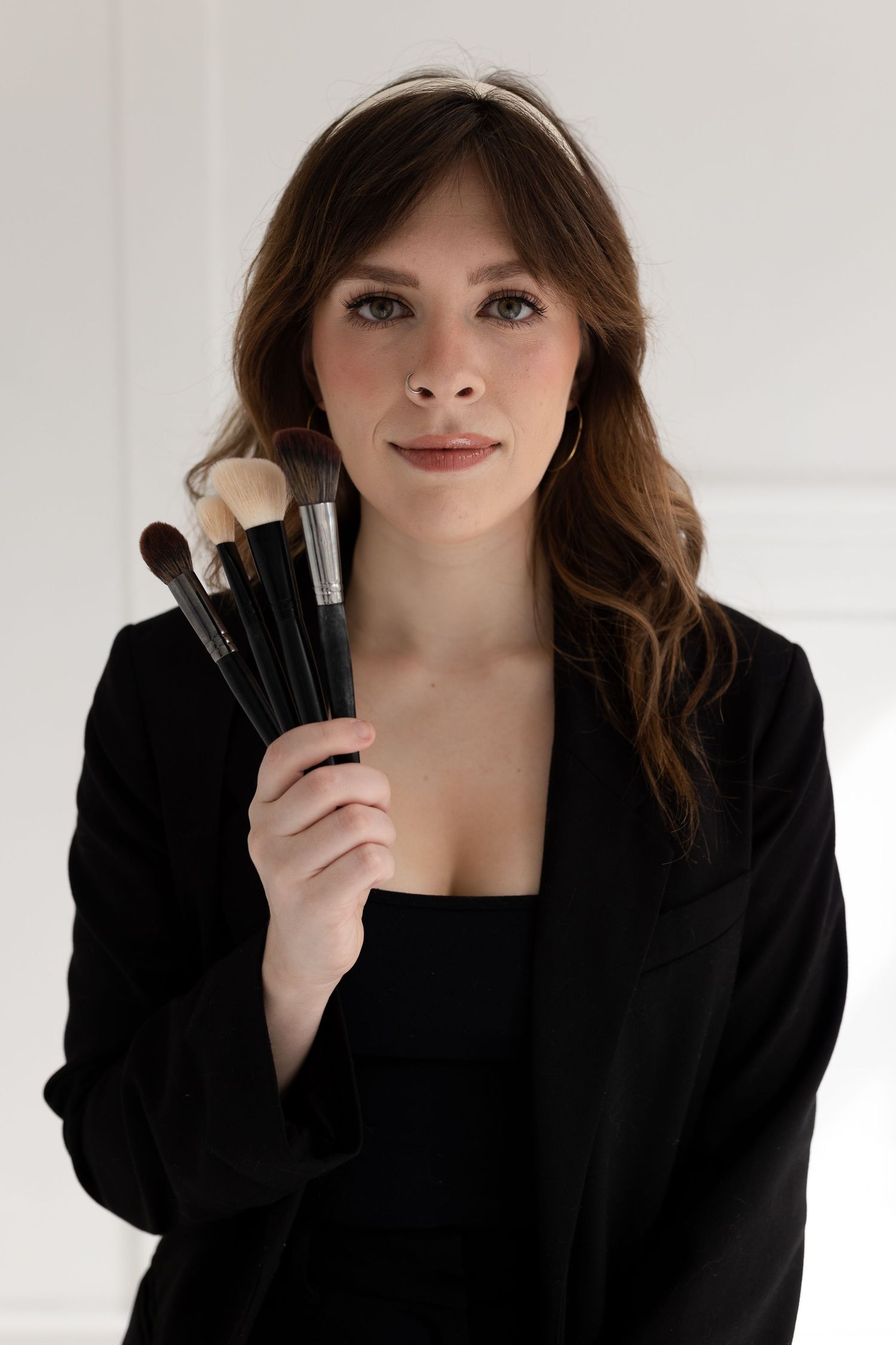 A person in a black blazer holding makeup brushes against a white background.