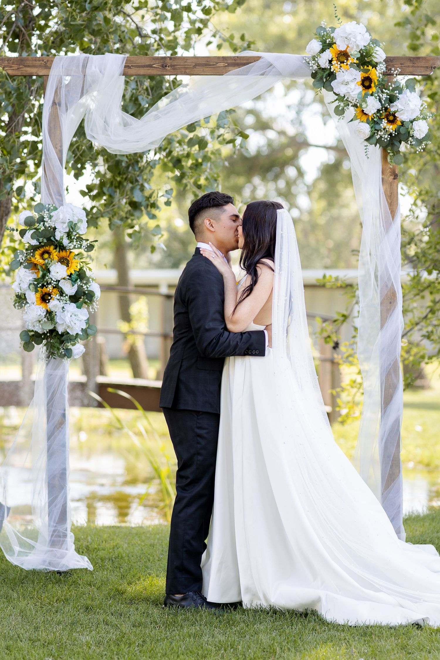 Wedding ceremony under decorated outdoor arbor with white fabric and sunflower arrangements.
