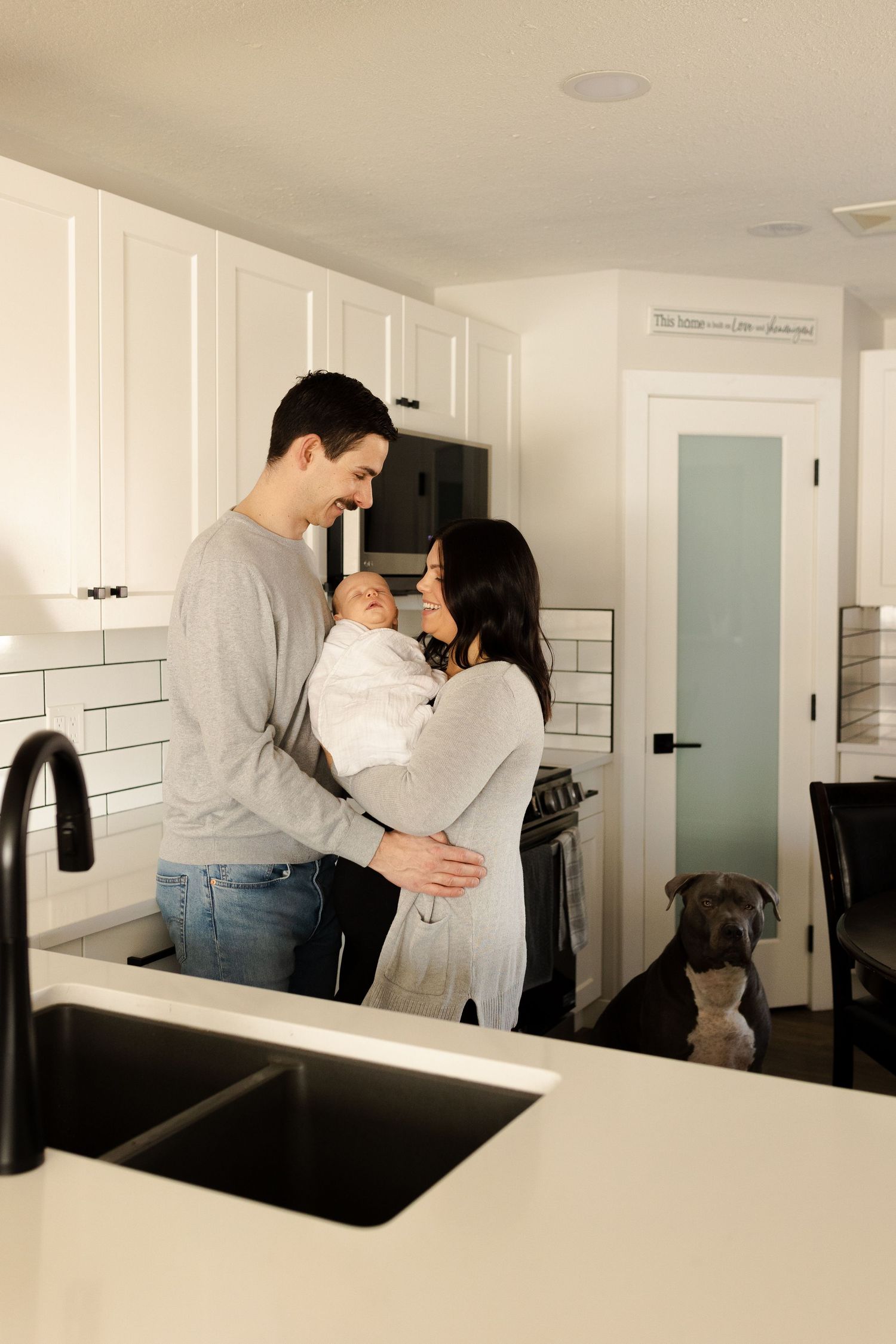 A couple stands in their modern white kitchen holding a baby while their dog looks on from the corner.