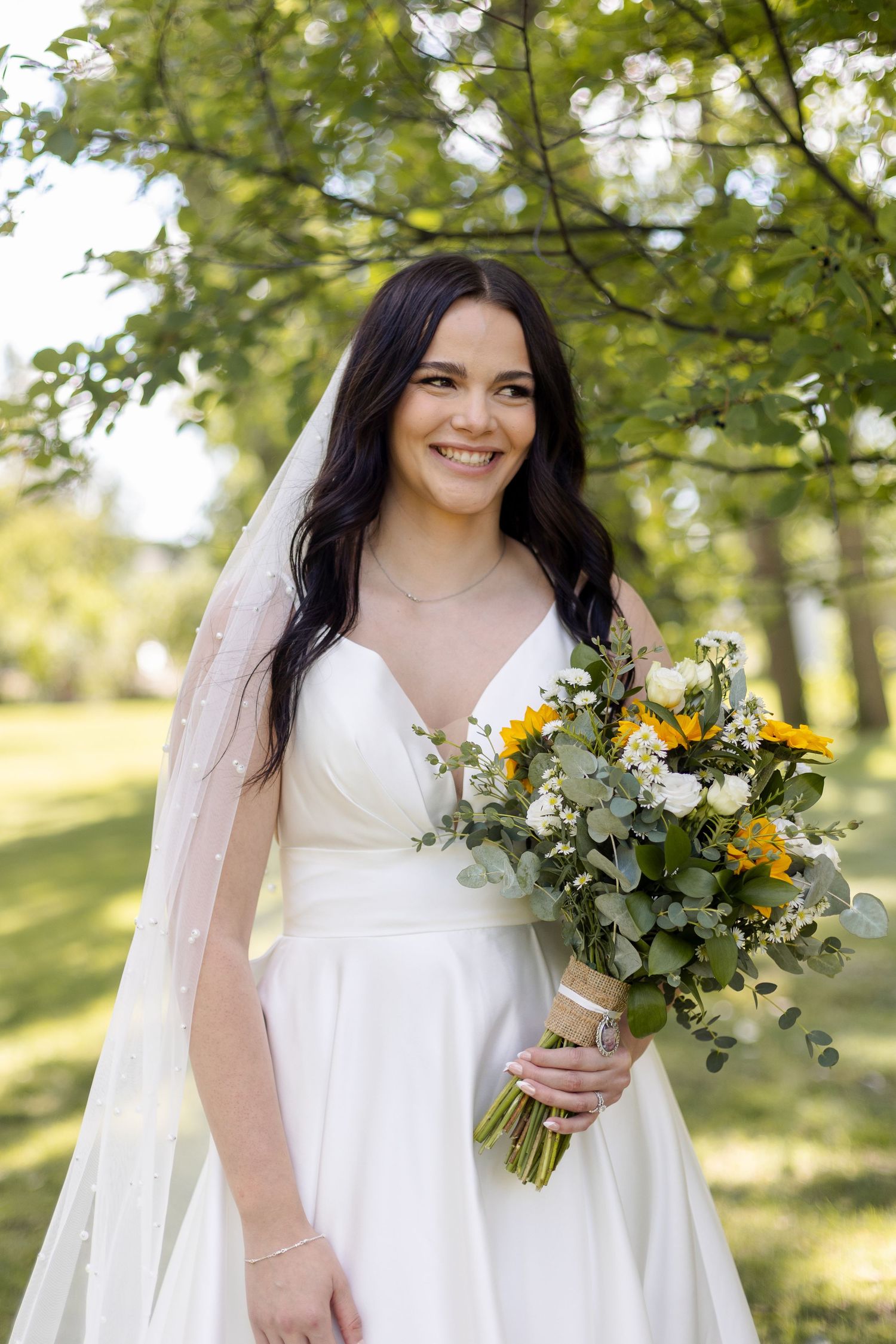 Bride in white dress holds yellow and green bouquet while smiling outdoors under trees.