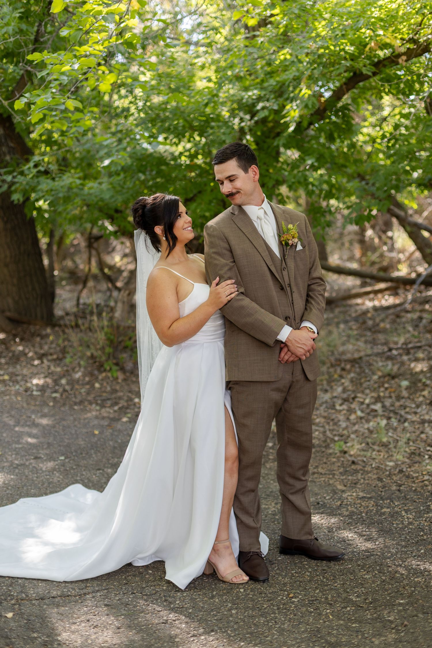 Wedding couple shares candid moments sitting together on ground in a wooded outdoor setting.