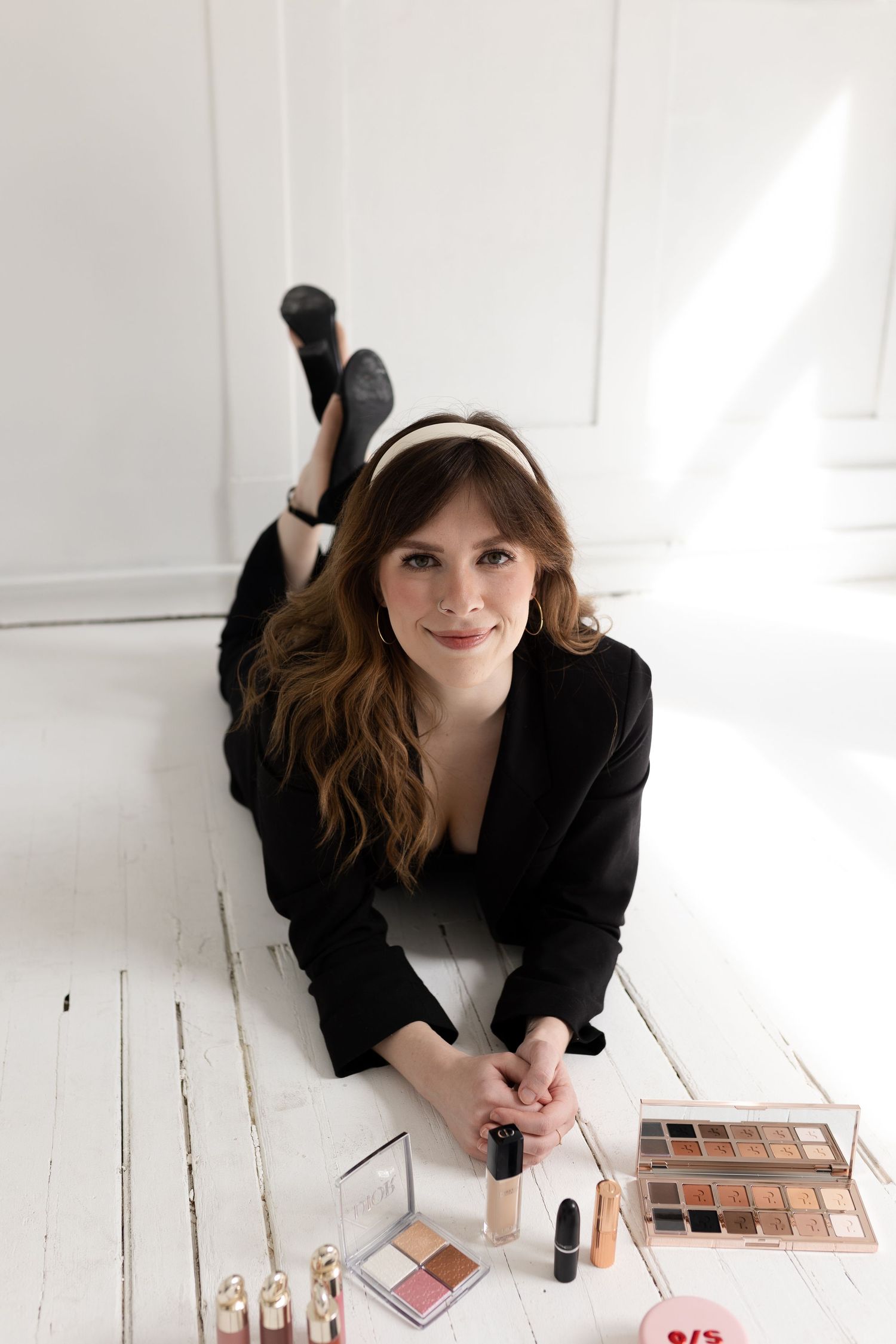 A person in black clothing lying on a white floor with makeup products nearby.