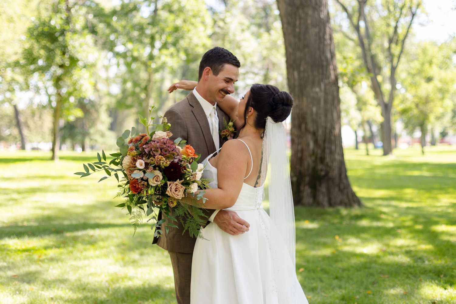 A romantic wedding couple embraces in a sunlit park surrounded by lush trees and autumn-colored floral bouquets.