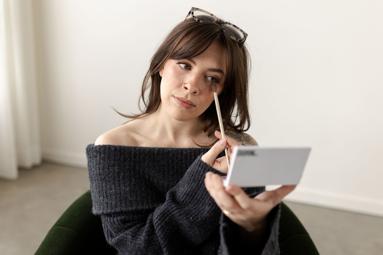Someone in a cozy gray sweater applies makeup while looking in a compact mirror.