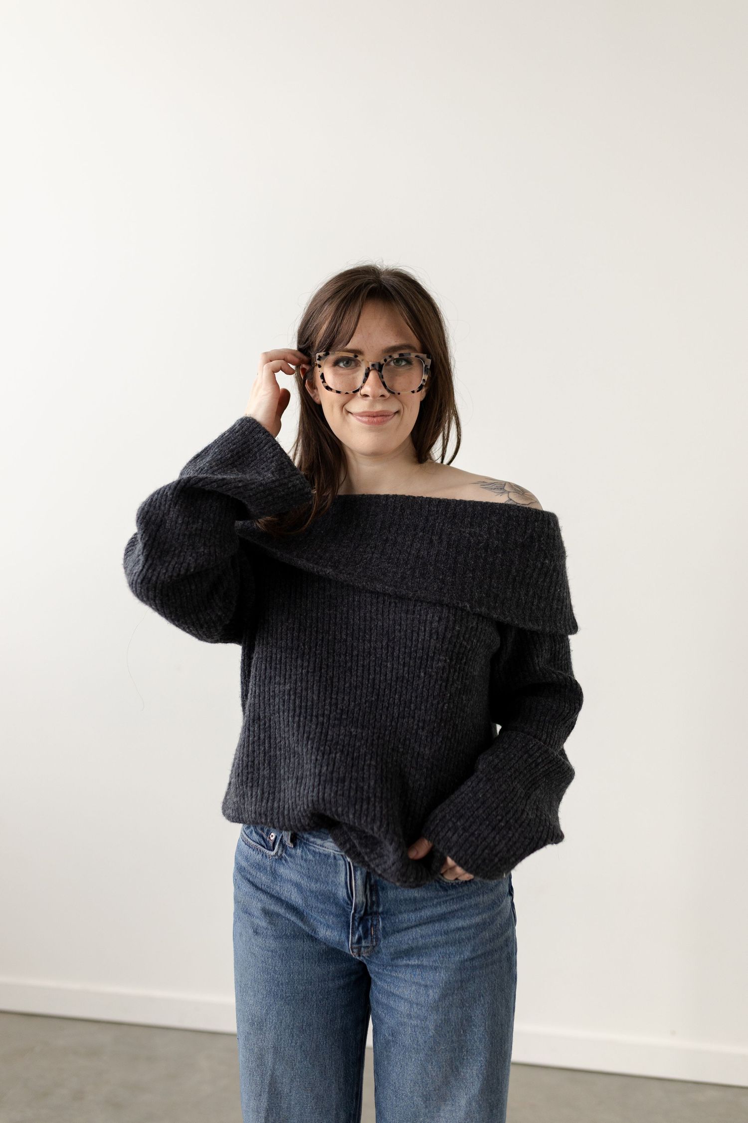 Person wearing an off-shoulder black sweater and jeans poses casually against a white wall.