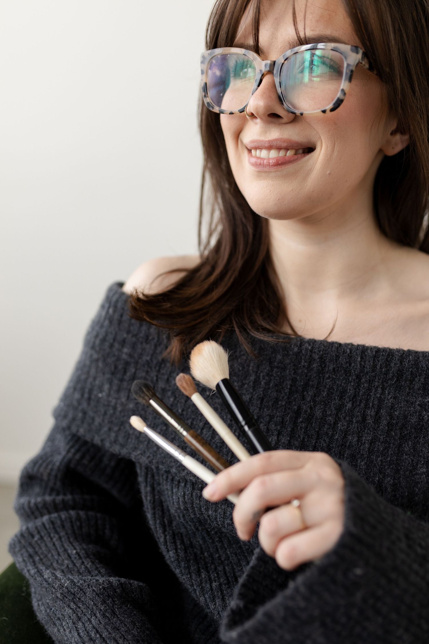 Person in black sweater and glasses holding makeup brushes while smiling against white background.