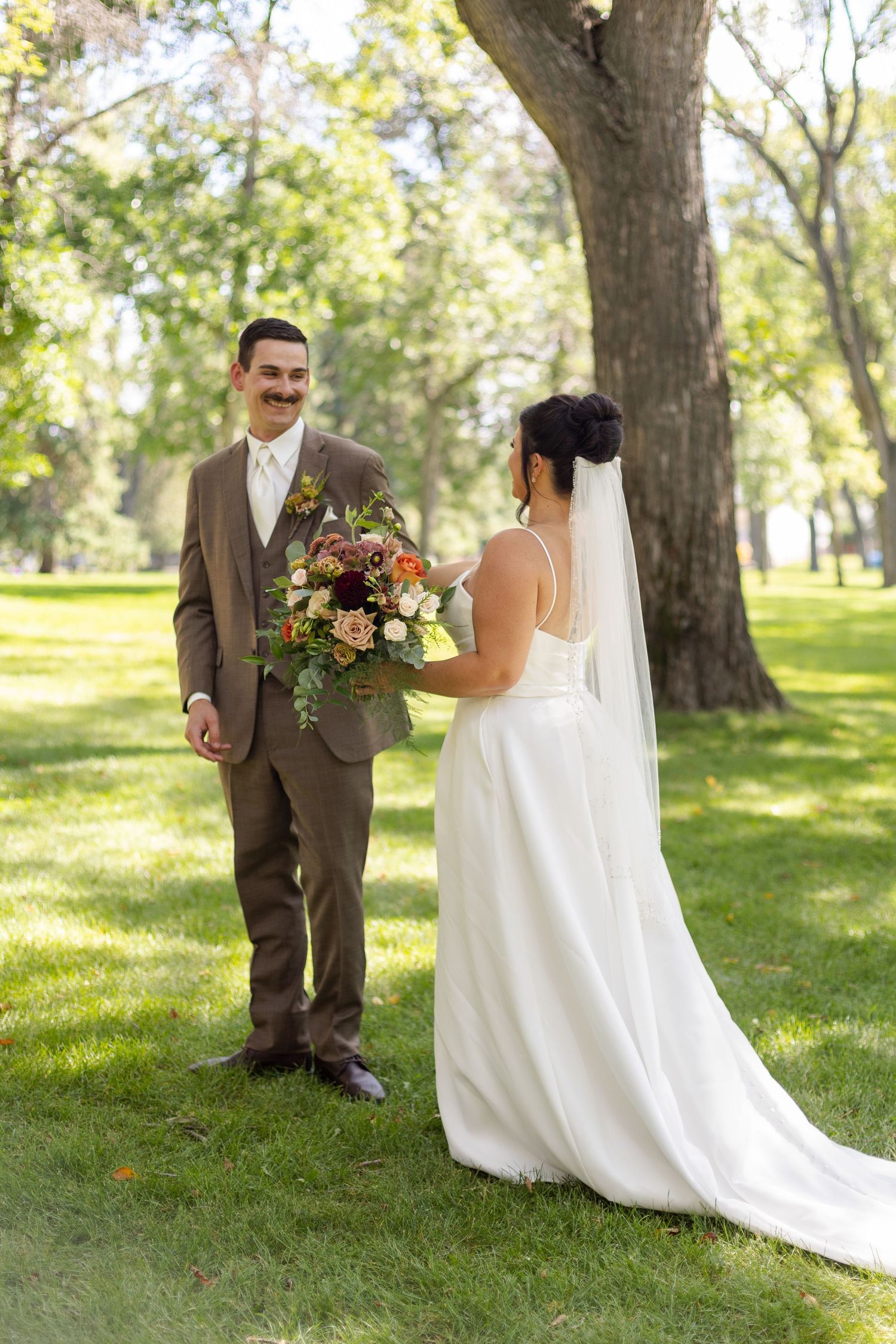 A couple shares a first look moment in a sunlit park with autumn trees and a beautiful bouquet.