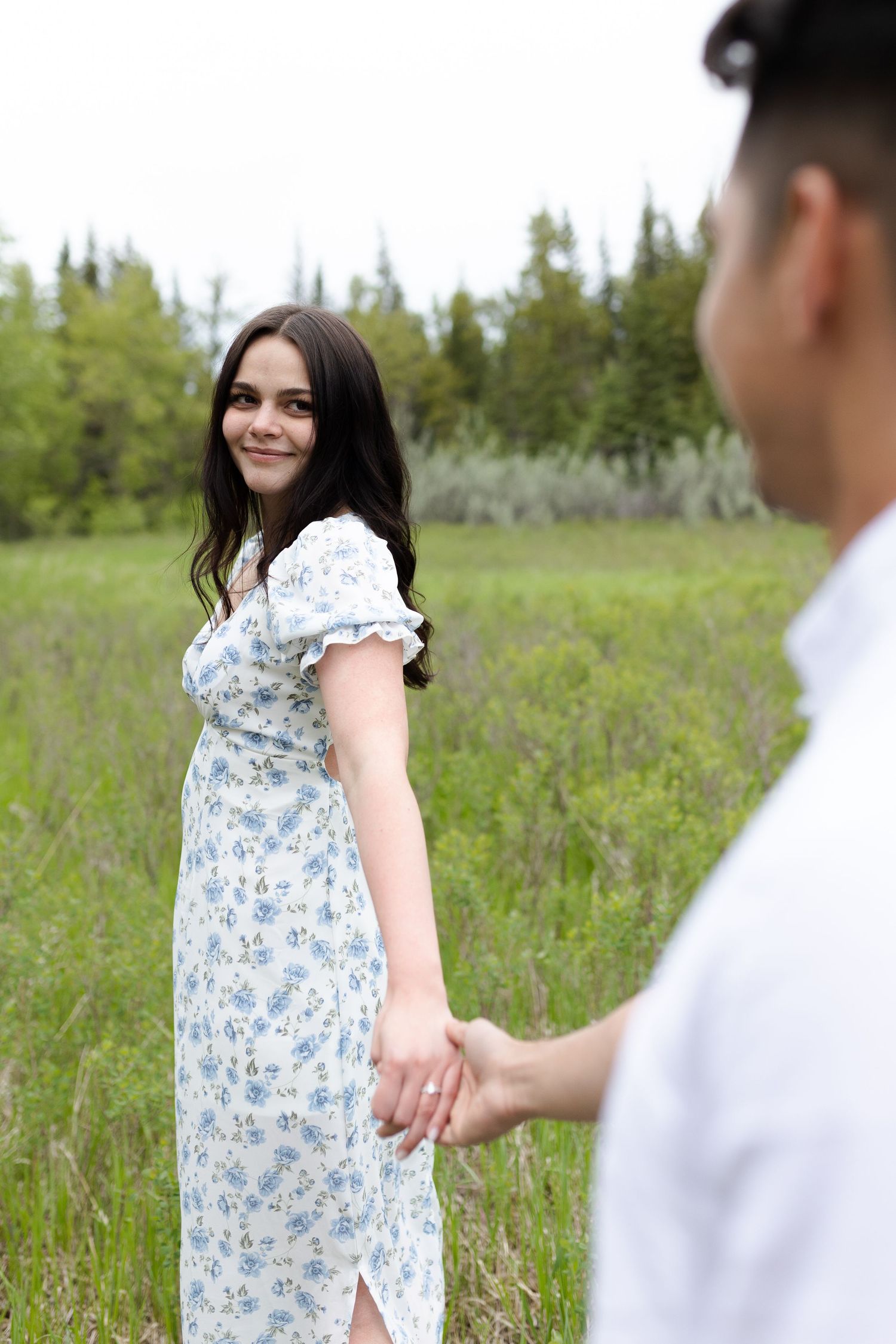 Couple in white and floral dress holding hands in a sunny meadow surrounded by pine trees.