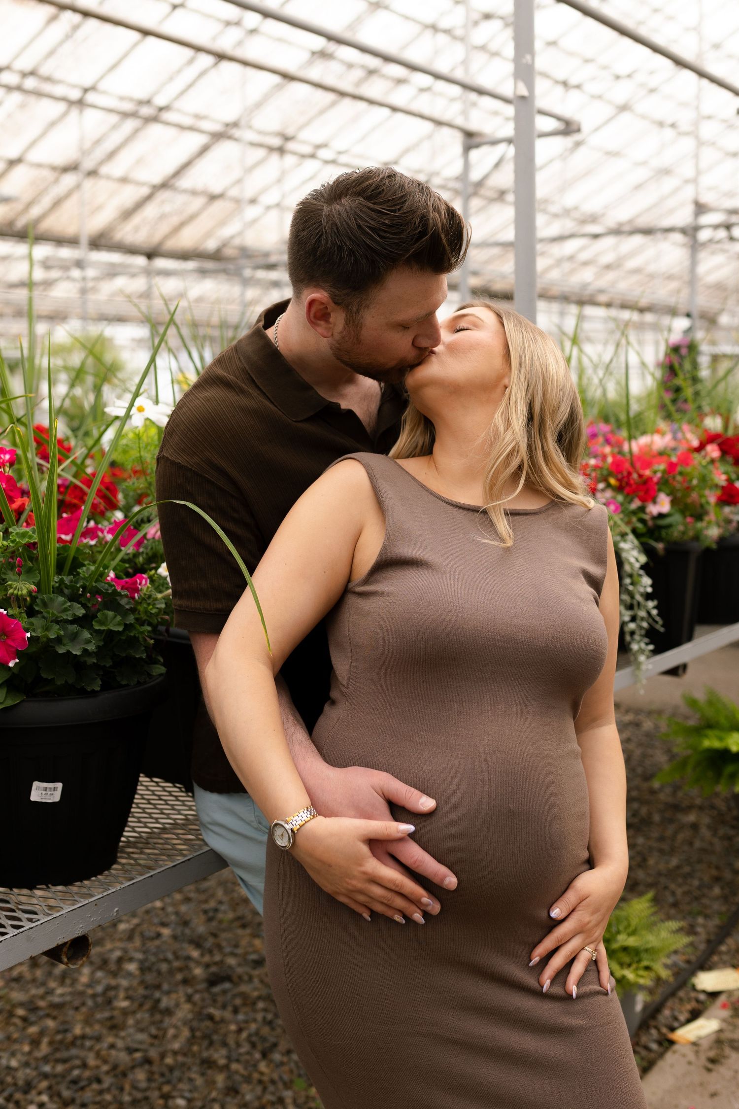 Intimate maternity photoshoot of couple embracing in a greenhouse with red flowers in the background.