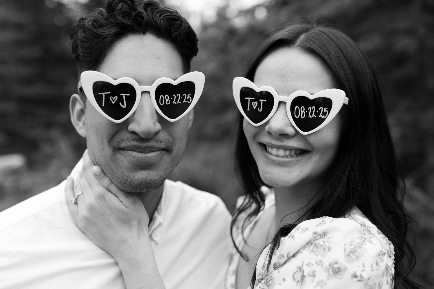 A playful couple wearing heart-shaped sunglasses poses for a black and white portrait.