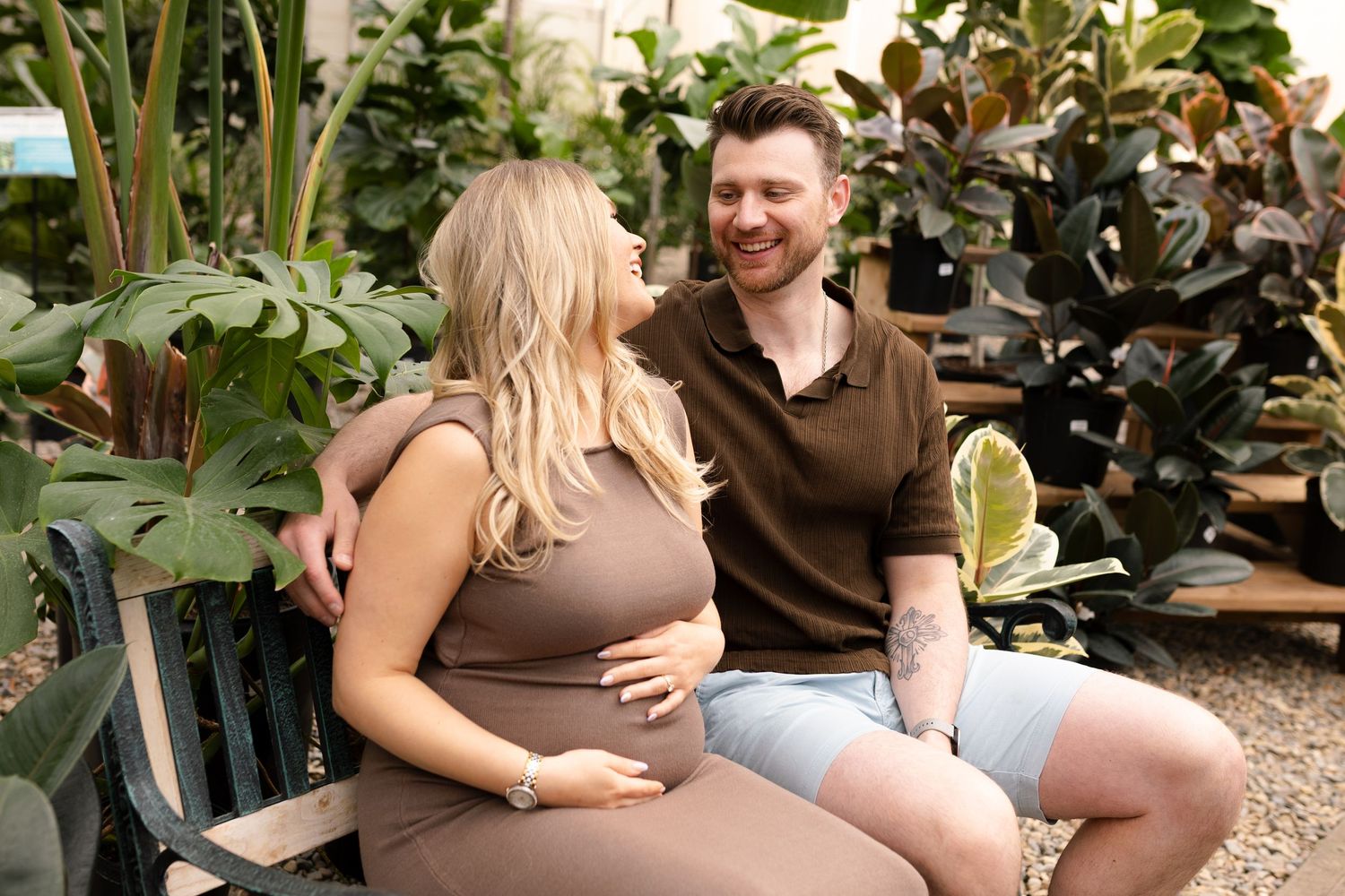 Candid maternity moment captured in greenhouse with couple sitting among tropical plants.