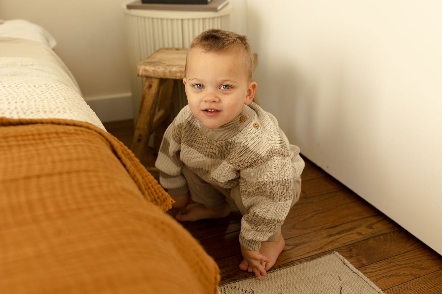 A baby in a cream sweater crawls on a bed with warm lighting and neutral tones.