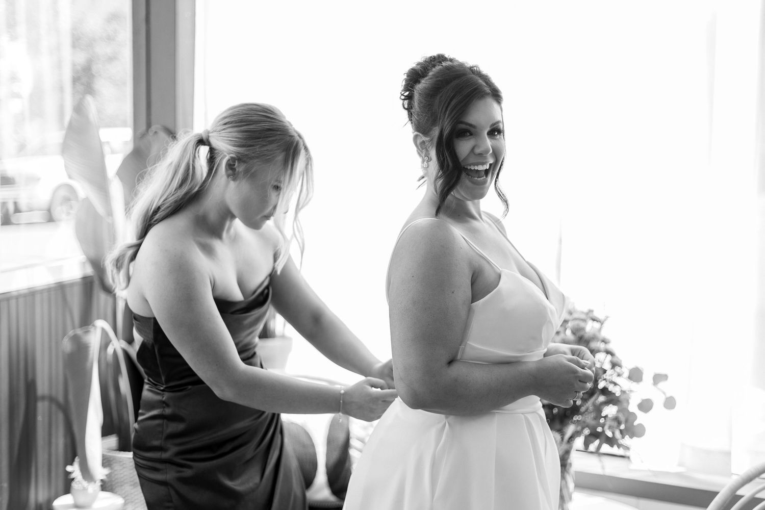 A bridesmaid helps adjust a wedding dress during final preparations in a sunlit room.