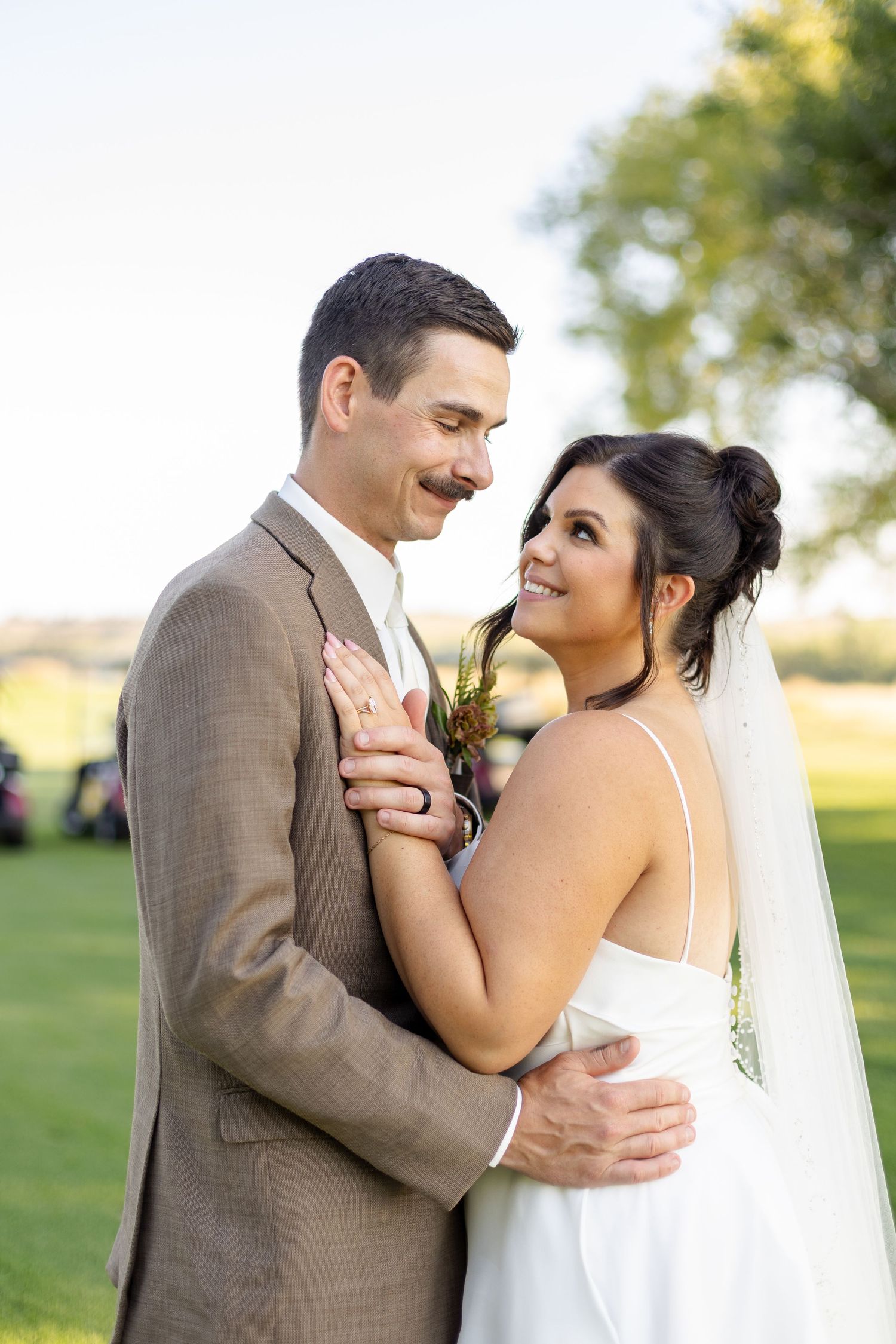 A bride and groom share an intimate moment outdoors on their wedding day, smiling at each other in formal attire.