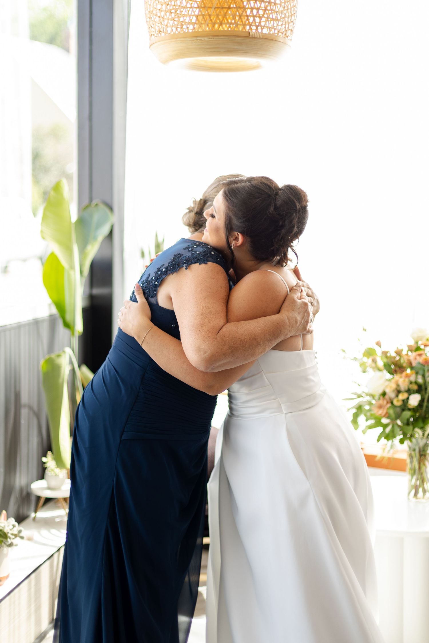 Two people share an emotional embrace at a wedding, one wearing a navy dress and one in a white bridal gown.