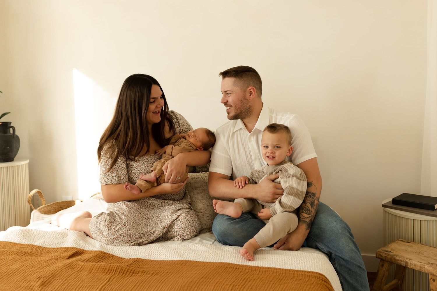 A family sits together on a bed in a cozy bedroom with warm lighting and neutral tones.