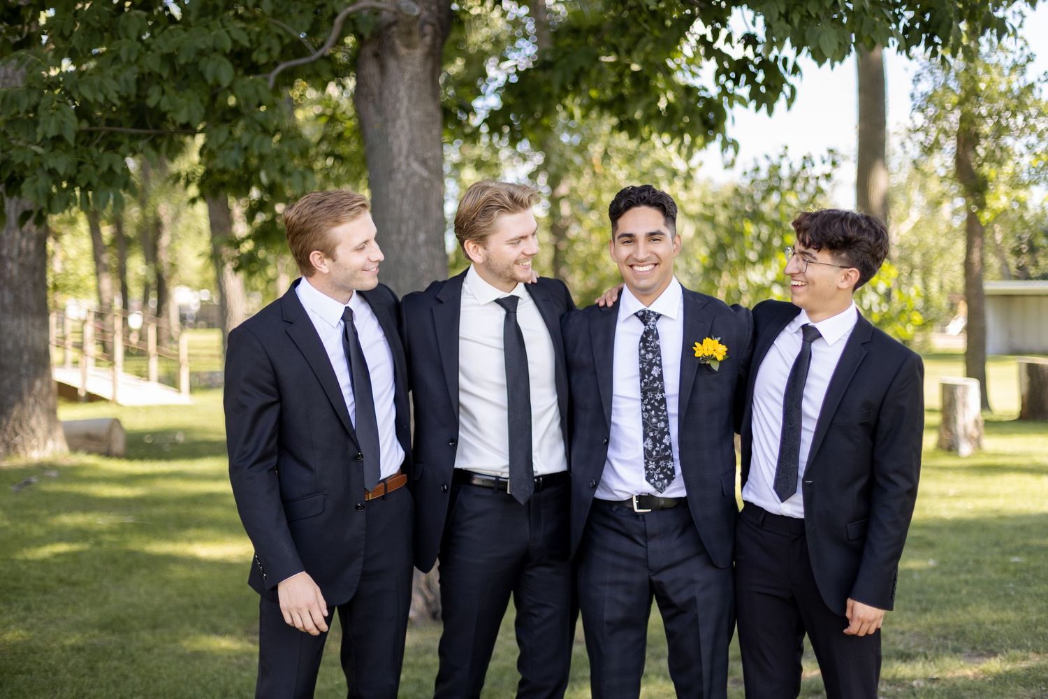Groomsmen in dark suits and ties pose together outdoors in a park setting.